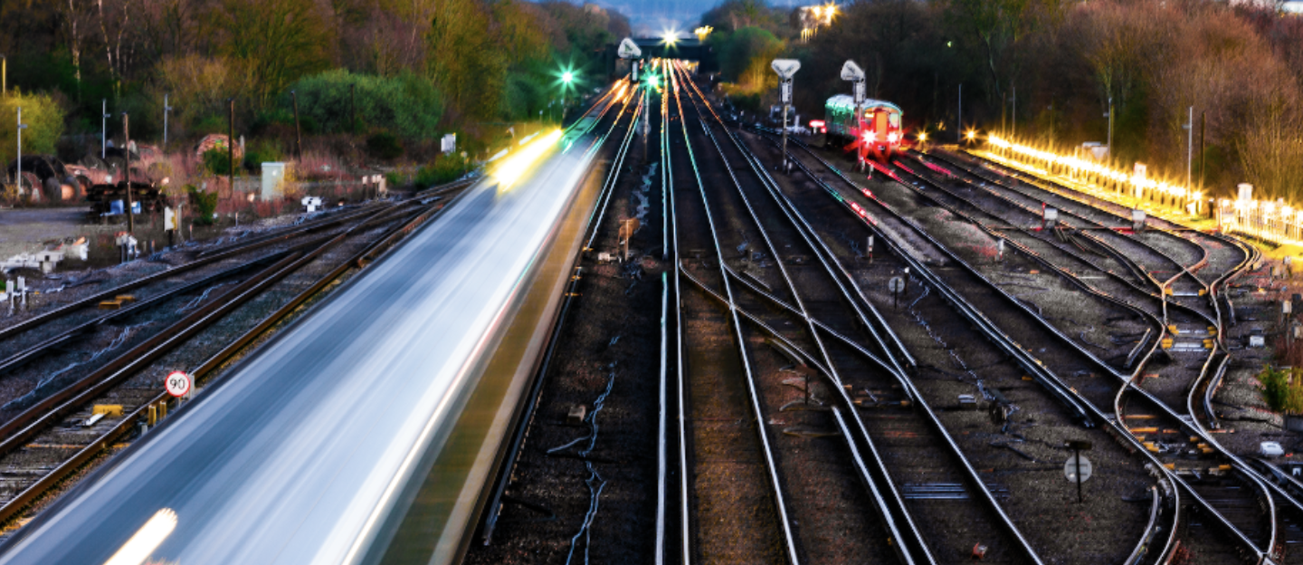 A view of railway tracks at dusk, with train lights creating streaks of light. Multiple tracks curve away into the distance, surrounded by trees and signaling lights.