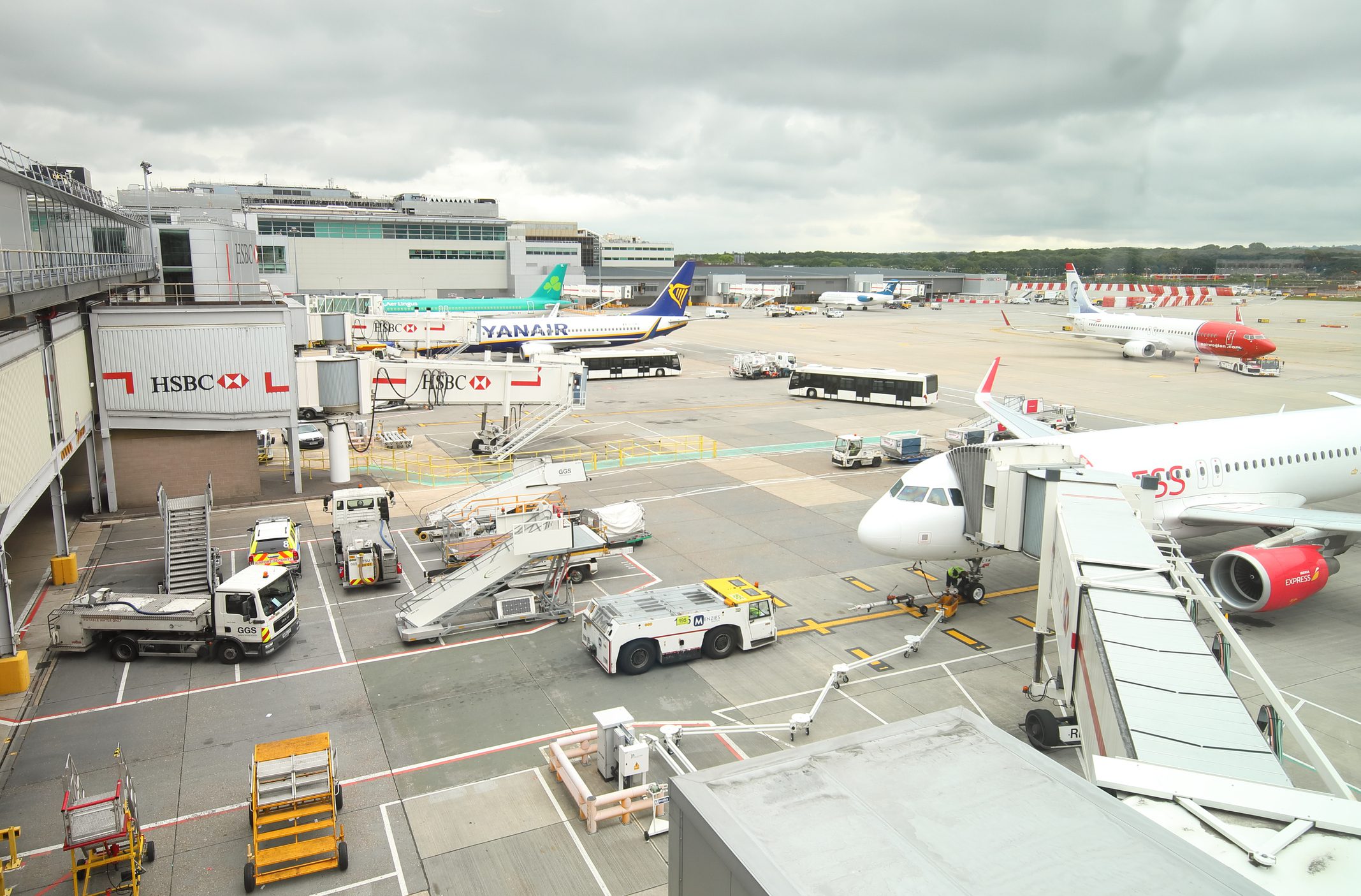 An airport scene showing multiple airplanes parked at gates, including a Yanair flight, and ground service vehicles assisting with luggage at a busy terminal.
