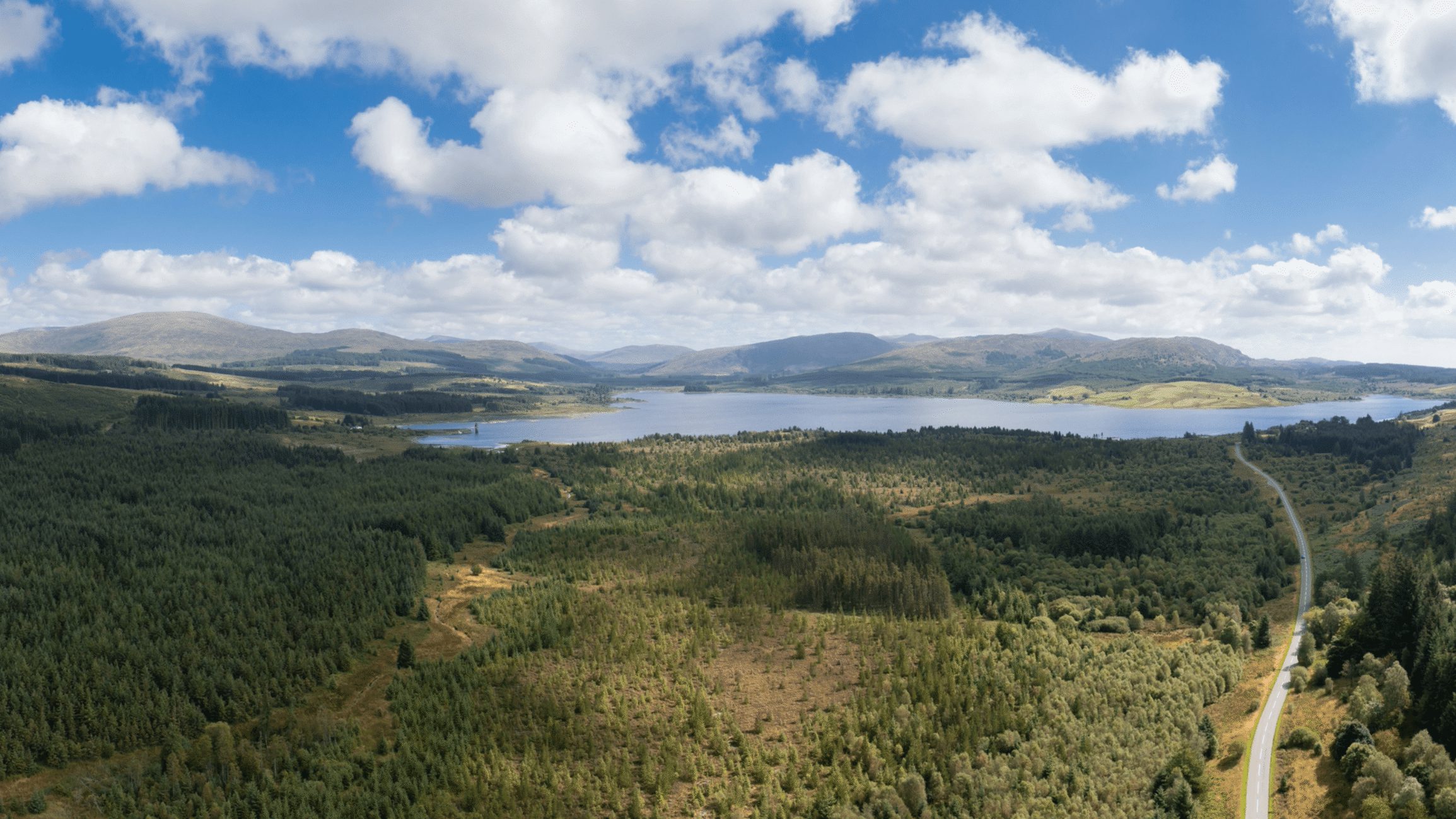 A panoramic view of a lake surrounded by green forests and rolling hills under a partly cloudy sky. A winding road is visible on the right side of the image.