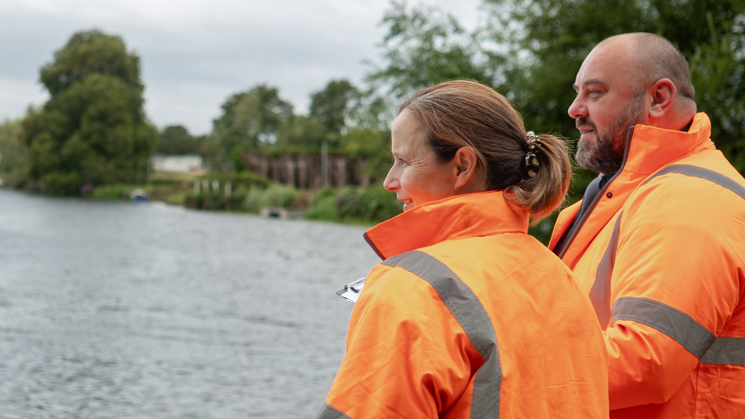 Two individuals in bright orange safety jackets standing by a riverbank, observing the water. The setting includes greenery and a cloudy sky.