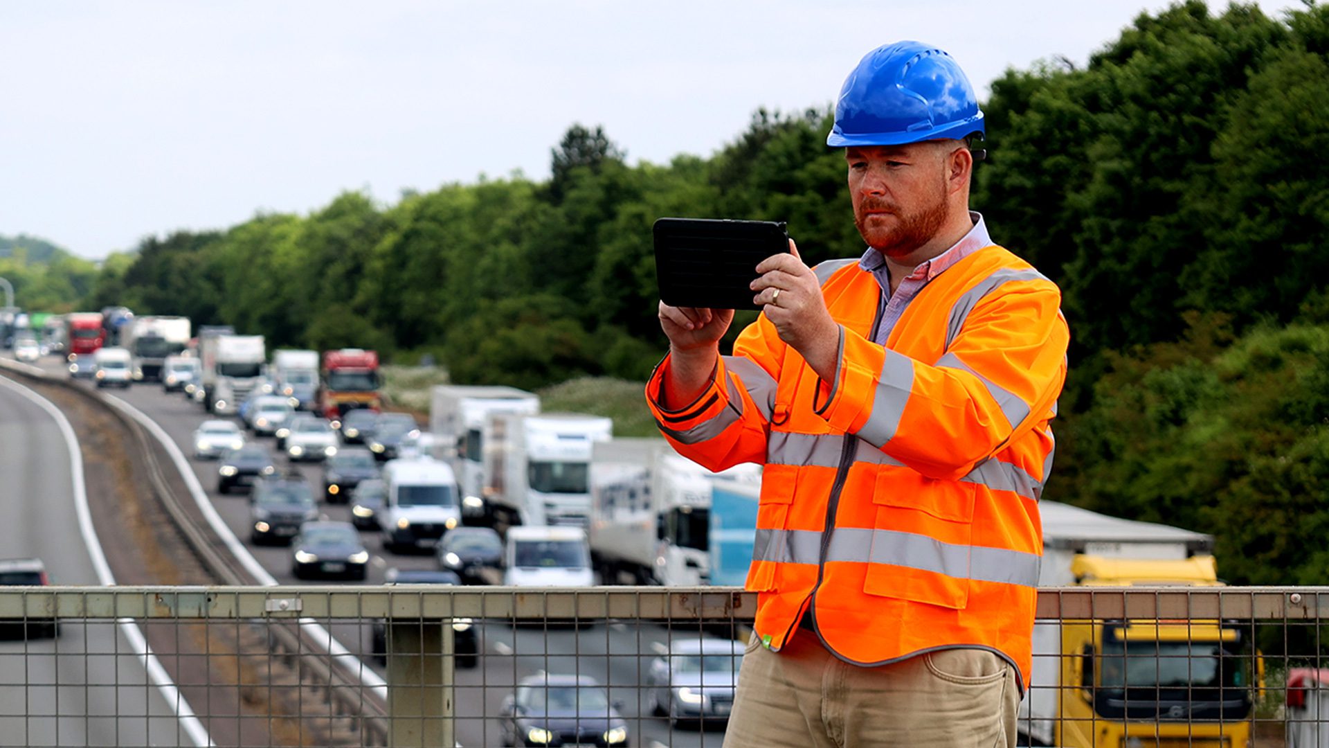 A construction worker in a bright orange safety vest and blue hard hat is using a tablet while standing on a bridge overlooking a busy highway filled with trucks and vehicles.
