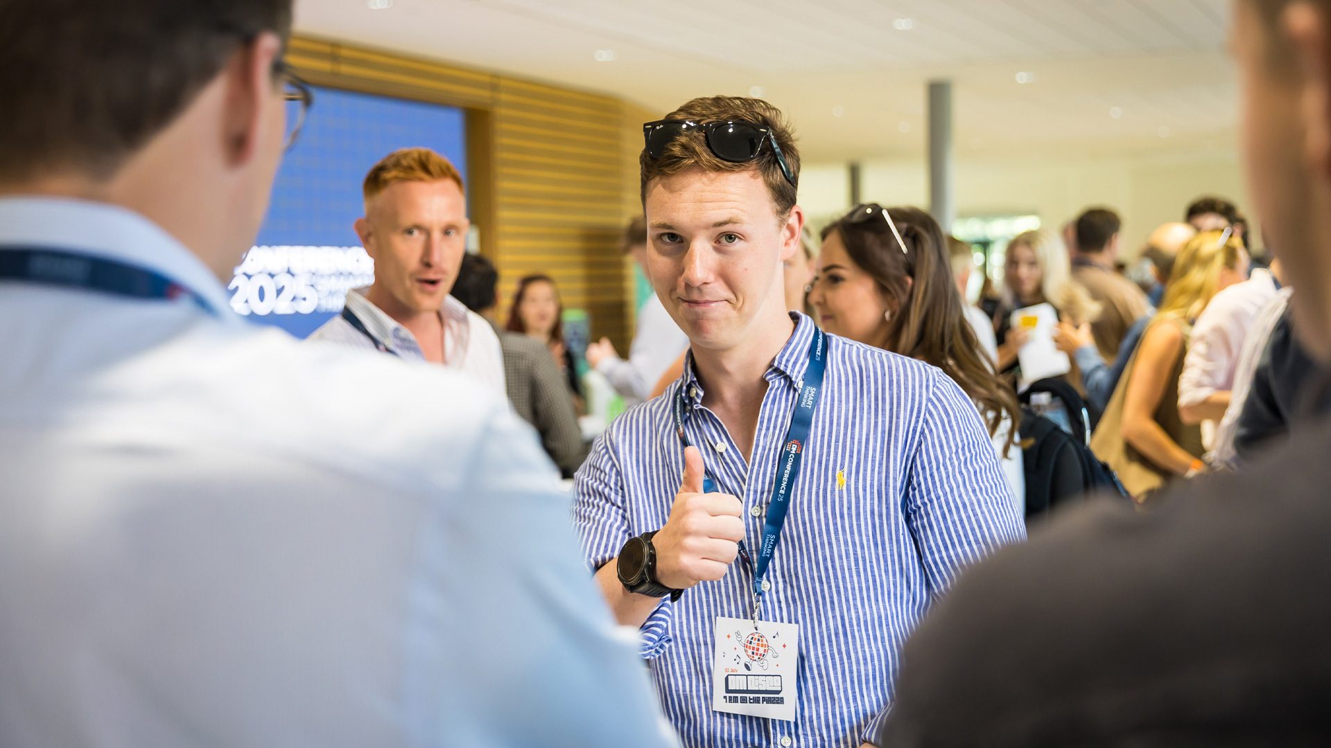 A young man giving a thumbs-up gesture during a networking event, wearing a blue striped shirt and lanyard. Others are engaged in conversation in the background, with modern decor visible.