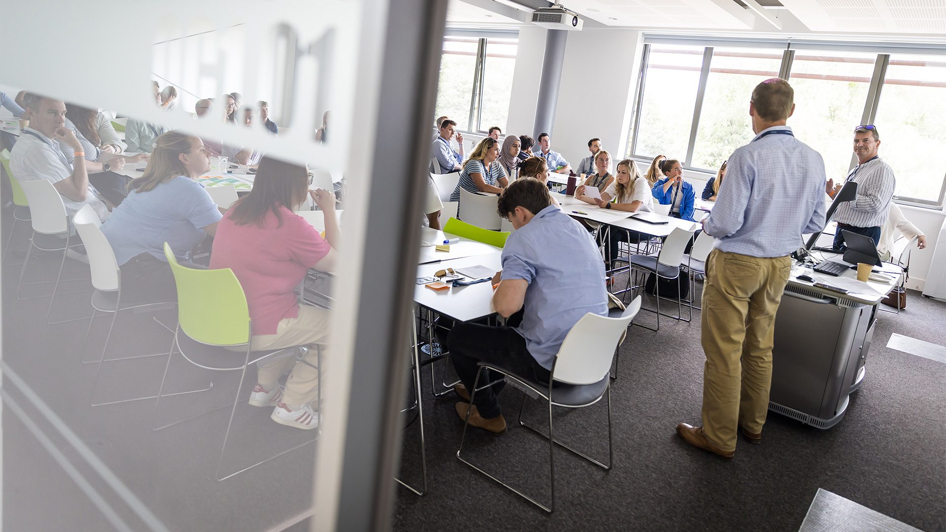 An office meeting with a speaker presenting to a large group of seated attendees. The room features modern furniture and is well-lit with large windows.