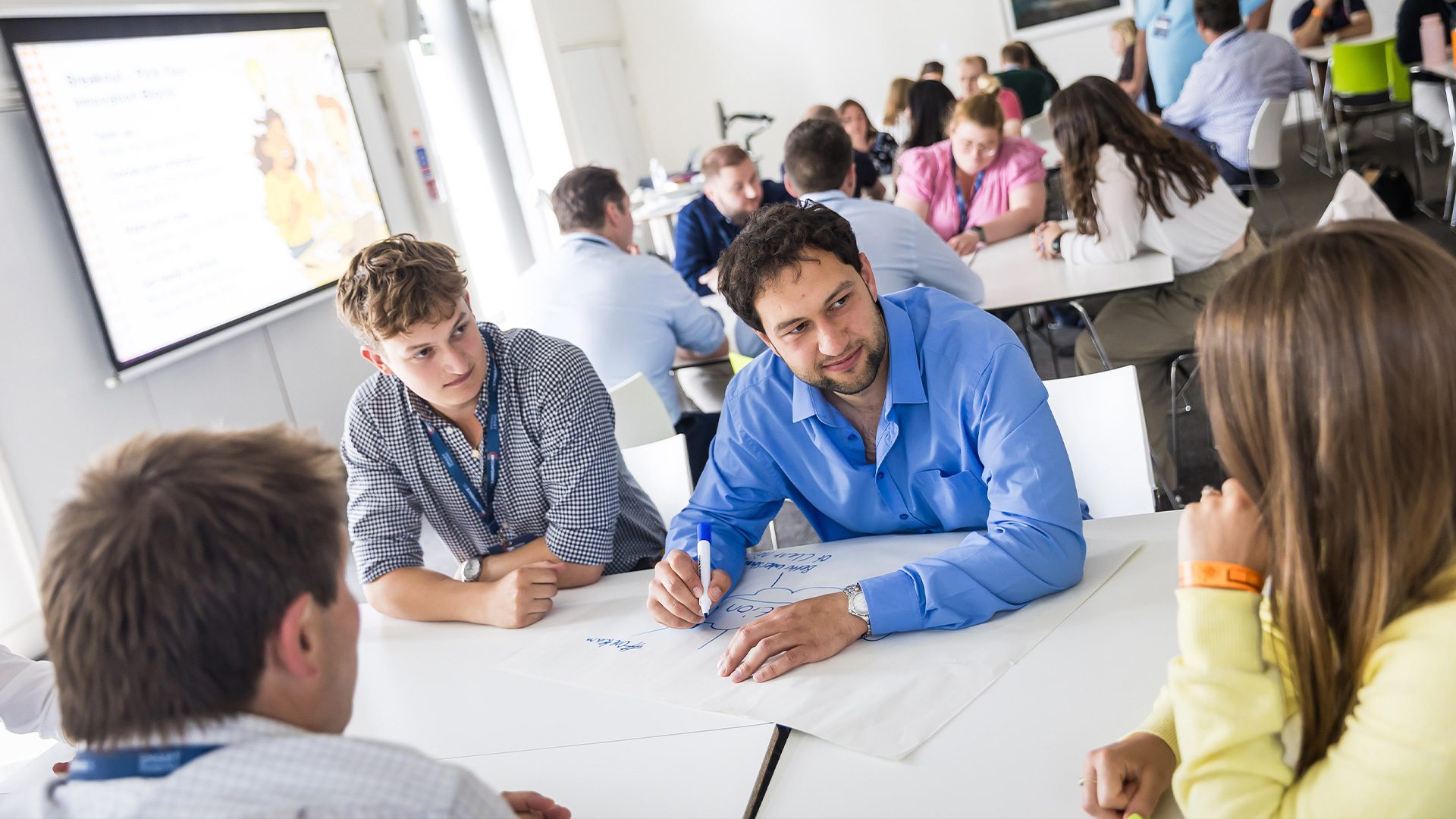 A group of people engaged in a collaborative brainstorming session at a table, with one person writing on a large sheet of paper. Others are observing and discussing ideas, while a presentation screen is visible in the background.