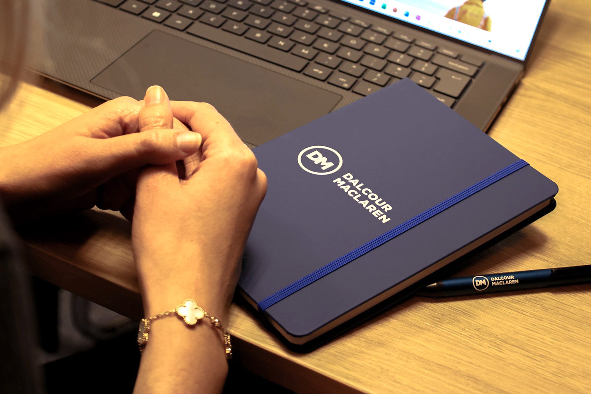 A person with a silver bracelet is resting their hands on a wooden desk near a closed notebook with a blue cover and the logo "DALCOUR MACLAREN." Next to it is a black pen and a laptop with an open keyboard.