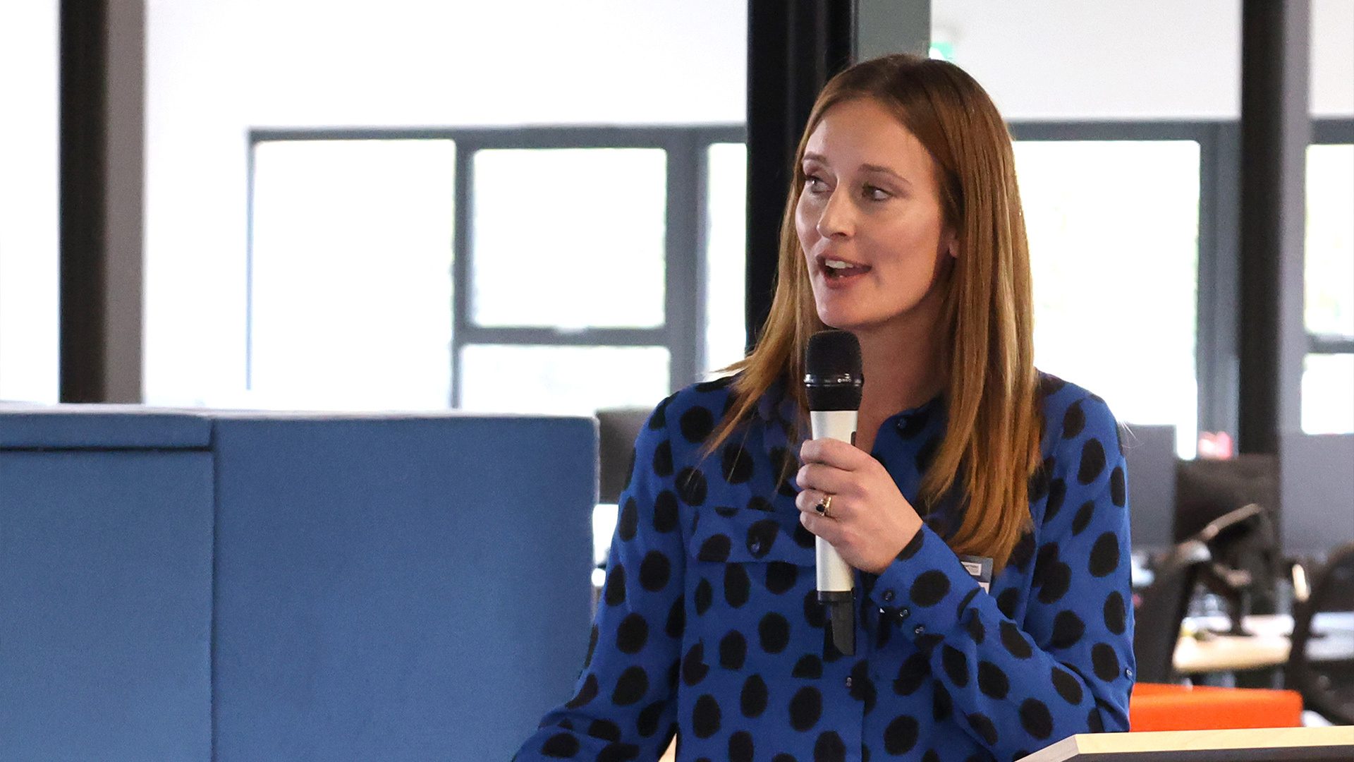 A woman with long brown hair wearing a blue blouse with black polka dots is speaking at a podium, holding a microphone, in a modern office setting with large windows in the background.