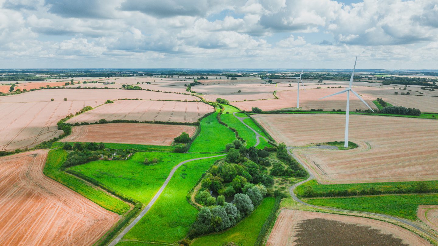 An aerial view of a rural landscape featuring fields of different shades, a winding green path, and a wind turbine in the distance under a partly cloudy sky.