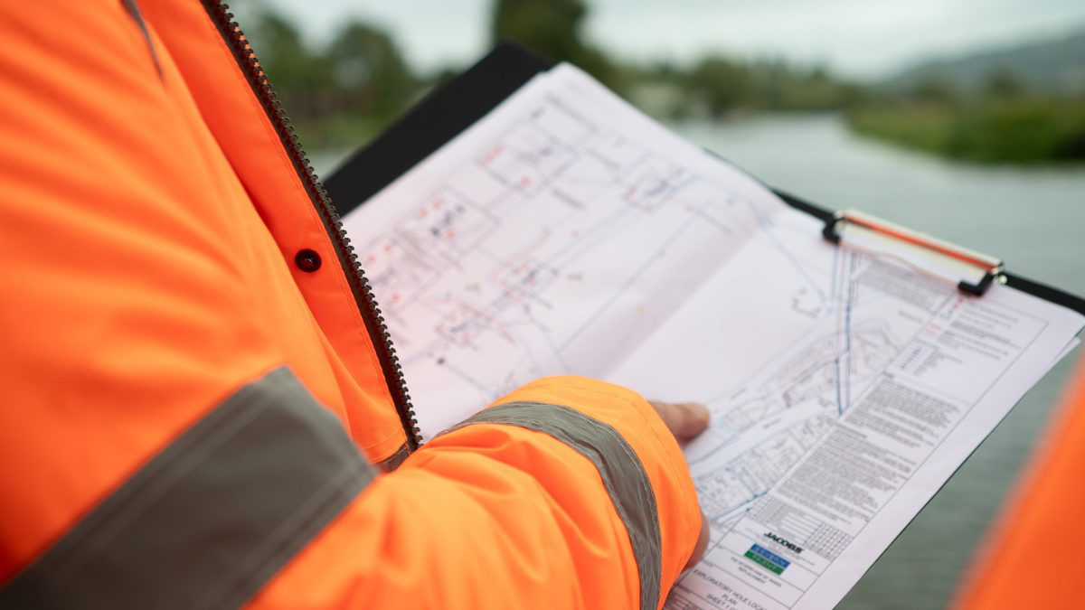 A person in a high-visibility orange jacket holding a clipboard with detailed plans, standing near a body of water in a natural setting.