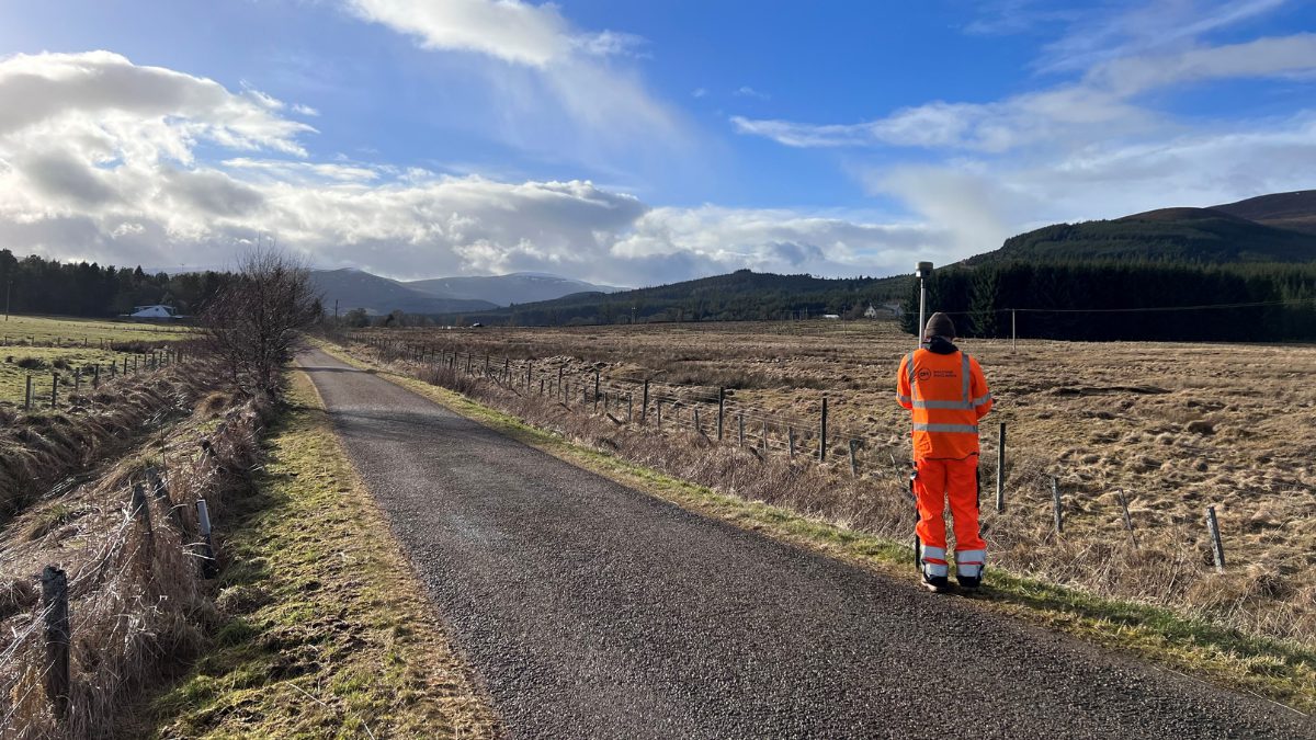 A person wearing an orange high-visibility jacket stands on a rural road, looking toward a scenic landscape of hills and fields under a partly cloudy sky.