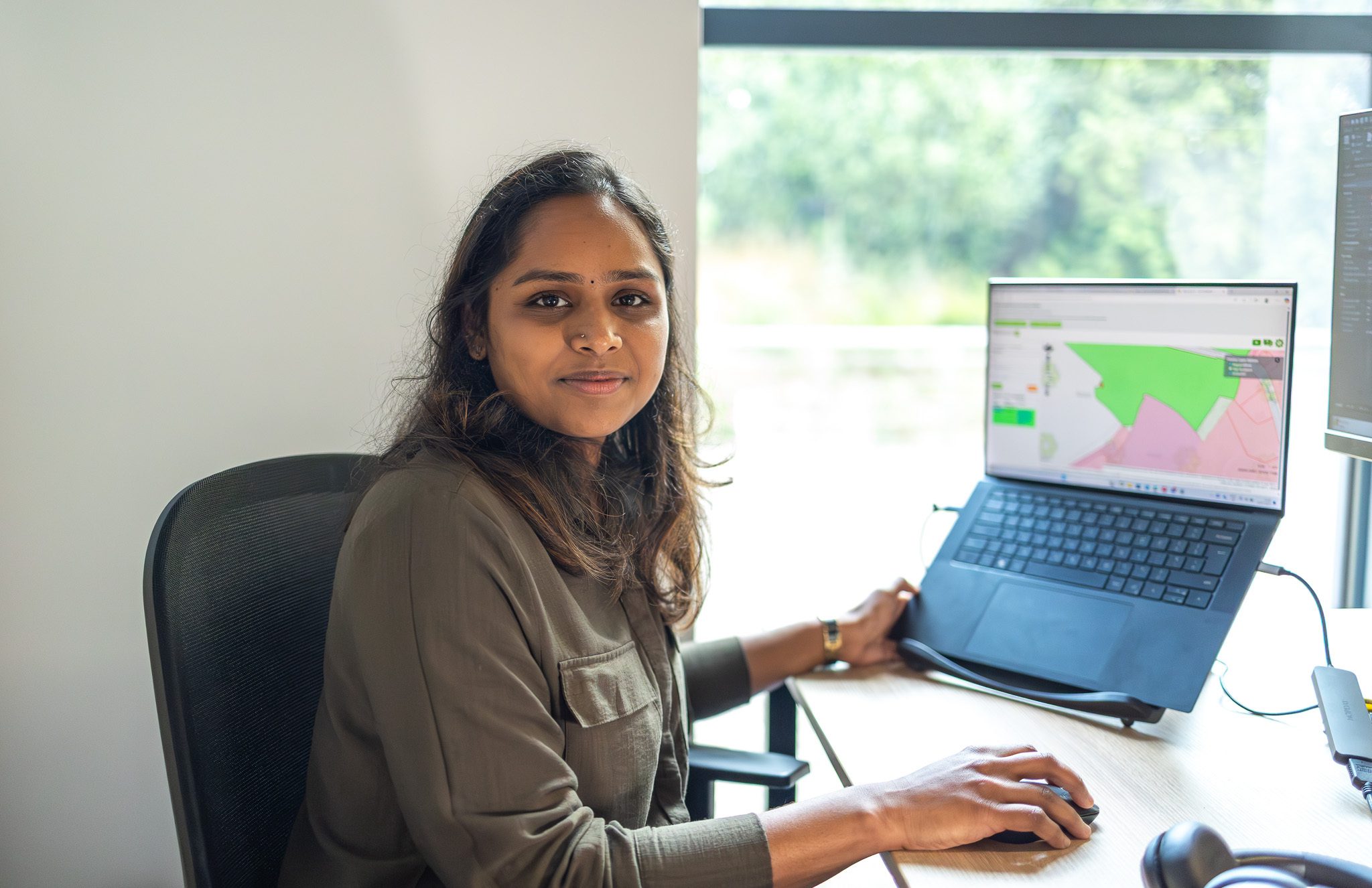 A woman sitting at a desk with a laptop, smiling while using a computer mouse. The laptop screen displays a colorful map. Natural light enters the room through a window, revealing greenery outside.
