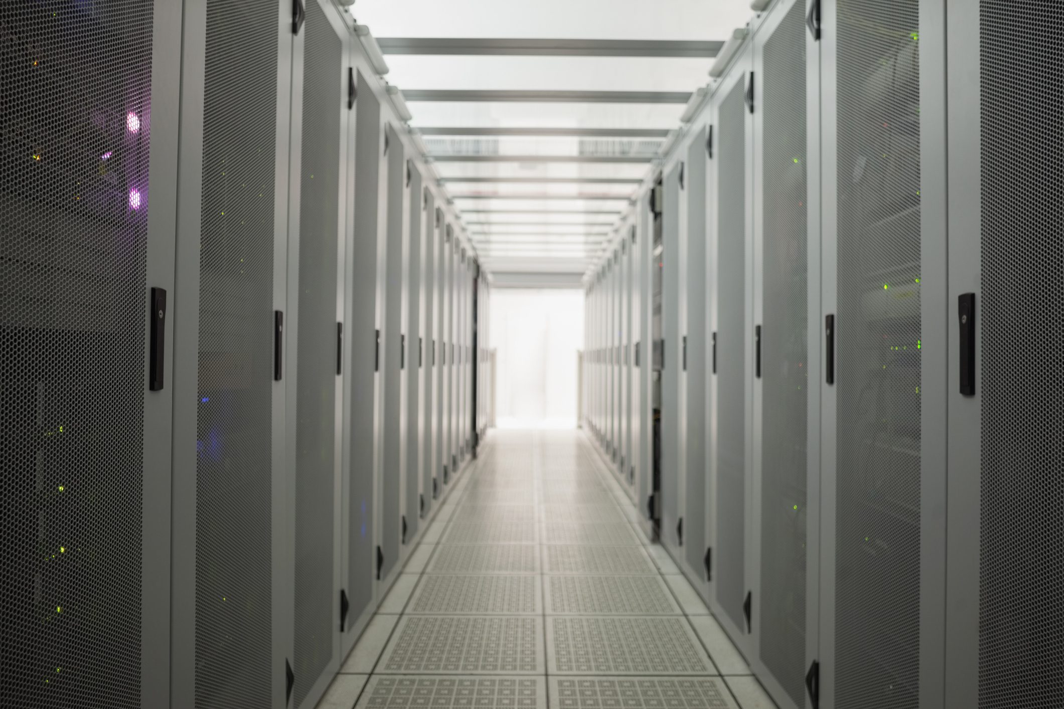 Interior of a data center featuring rows of server racks with a narrow aisle in between, illuminated by white lights at the far end.