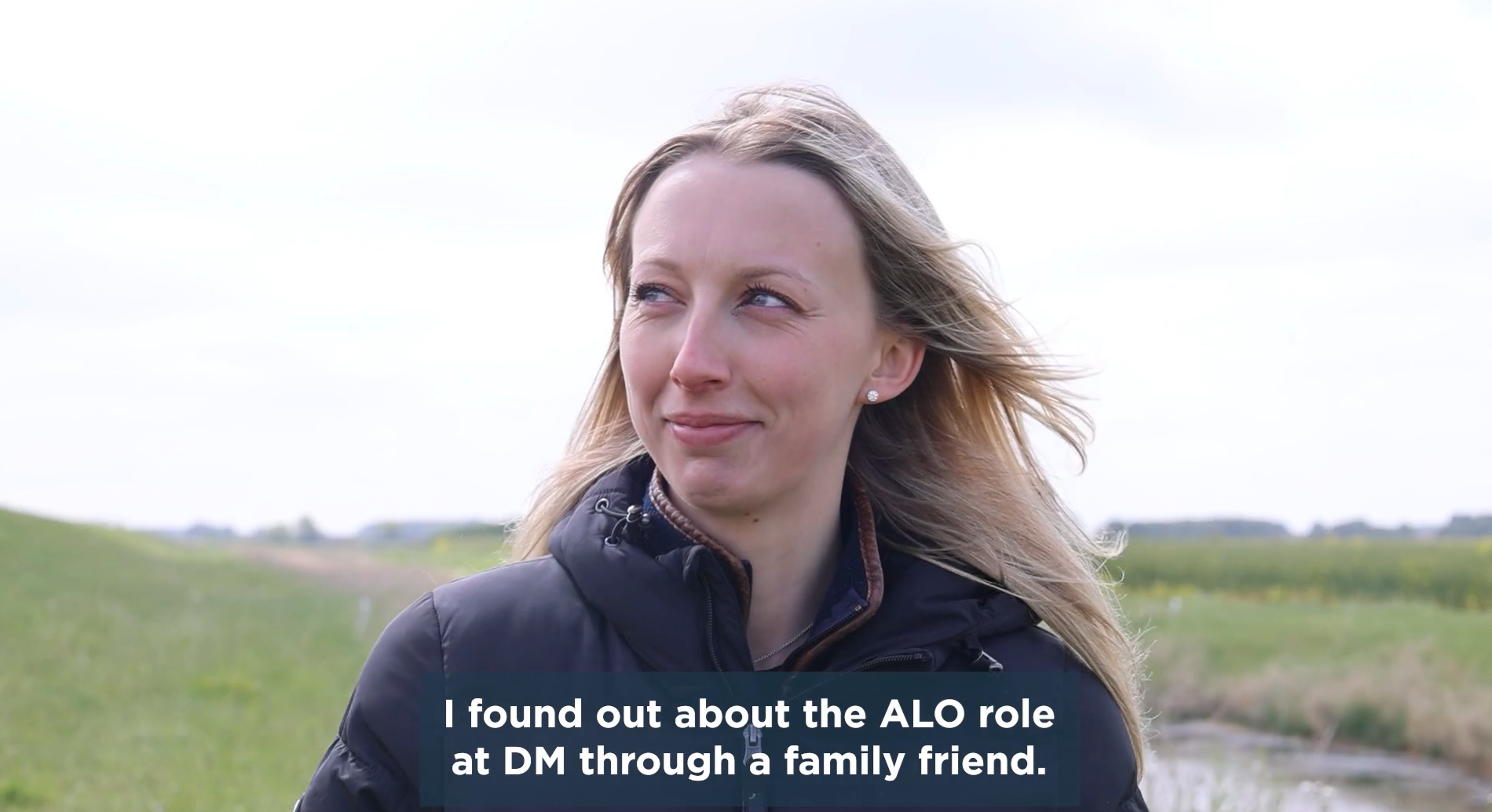 A woman with long, blonde hair standing outdoors, wearing a dark jacket, with green fields and a waterway in the background. Text on the image states she learned about an ALO role at DM through a family friend.