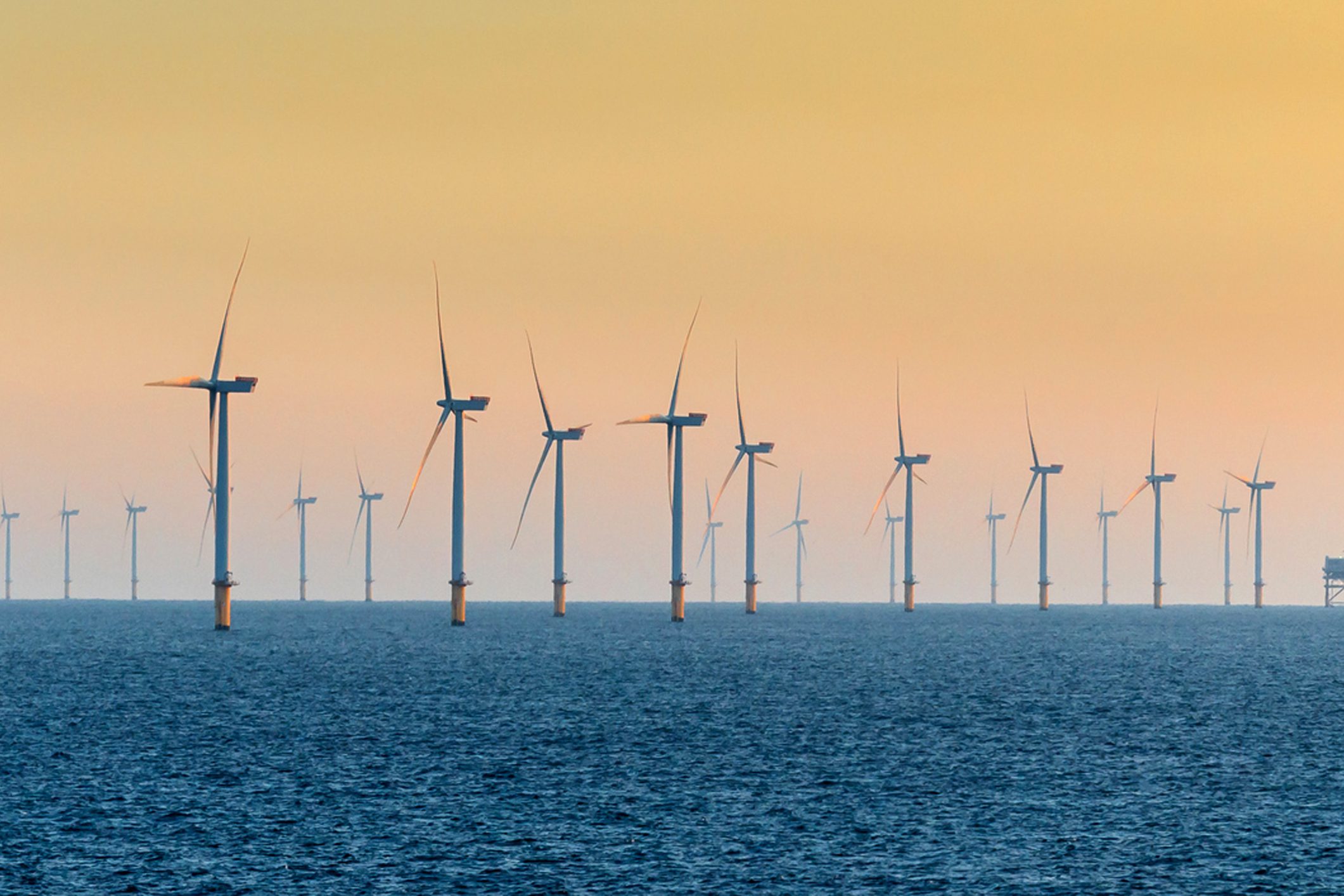 A series of wind turbines lined up in the ocean, with the sun setting in the background, creating a gradient of colors in the sky. The water is calm, reflecting the turbines.