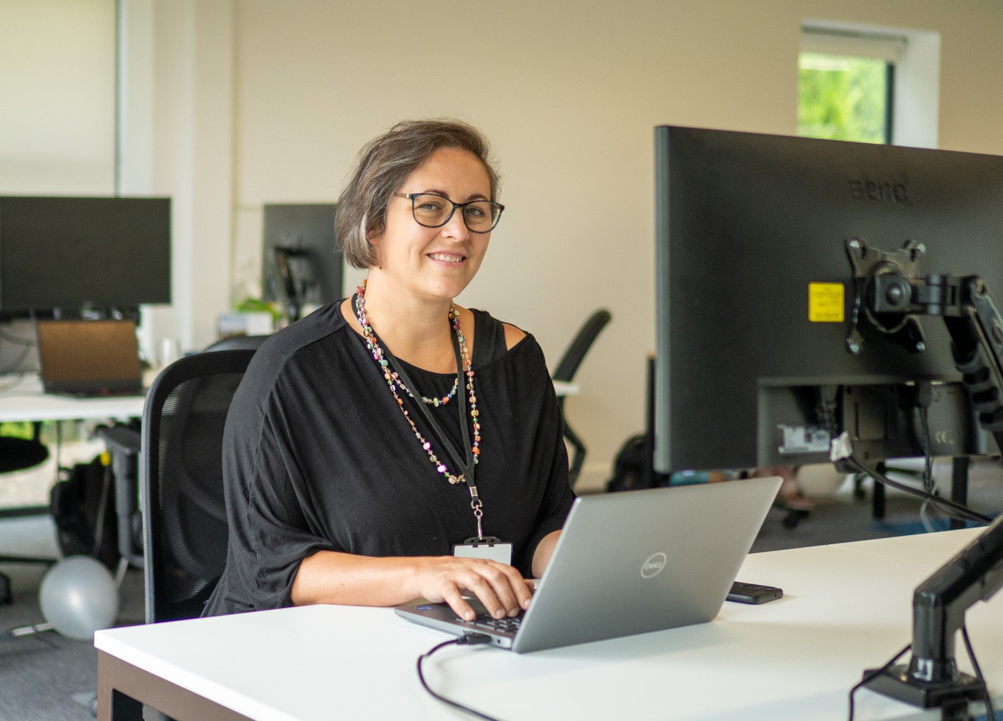 A woman with glasses seated at a desk, working on a laptop. She is wearing a black blouse and a colorful necklace, with office equipment and plants visible in the background.