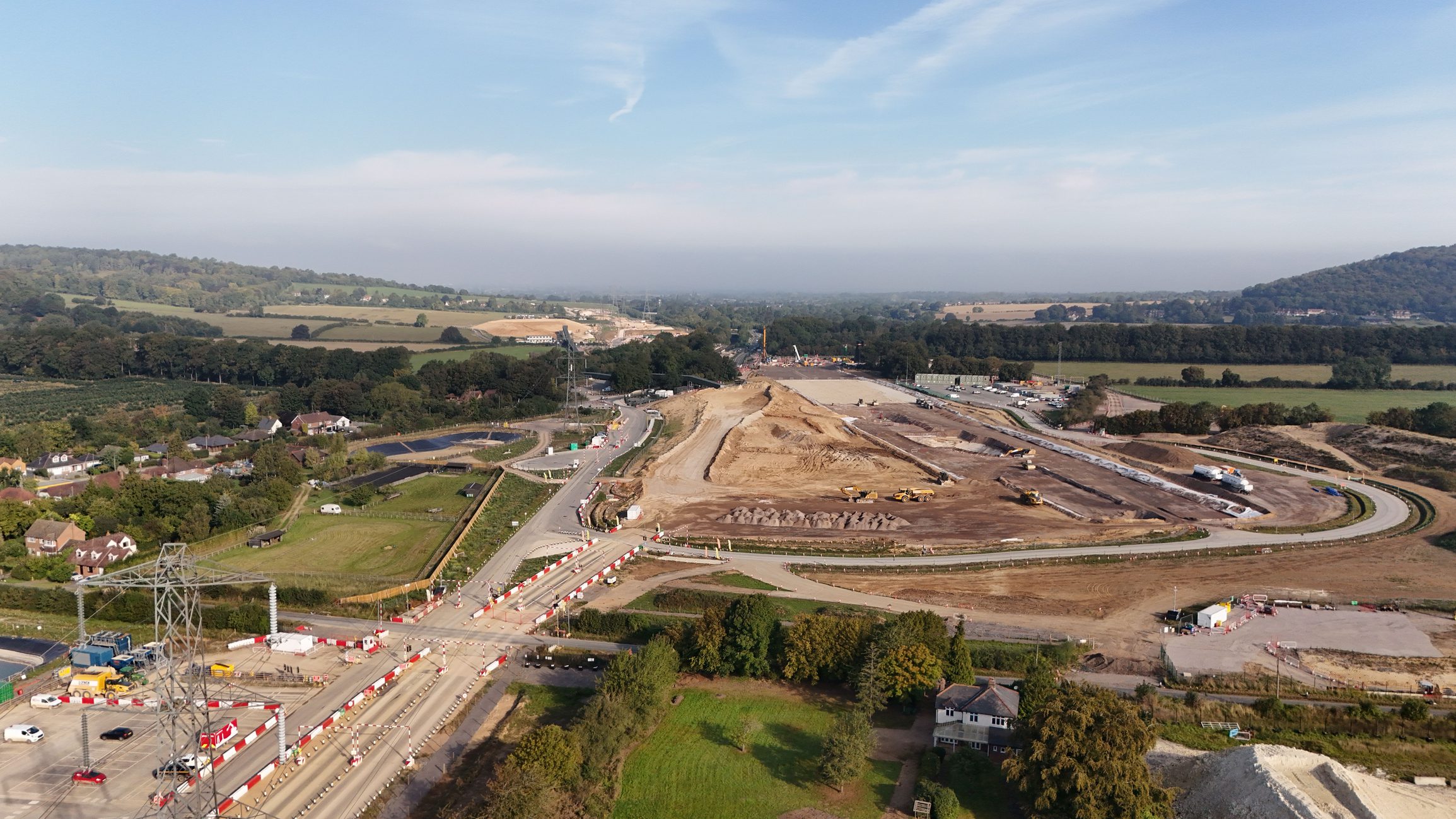 Aerial view of a construction site in a rural area, featuring large earth-moving equipment and cleared land. Surrounding fields and a distant wooded area are visible, with roads and traffic cones indicating ongoing work in the vicinity.