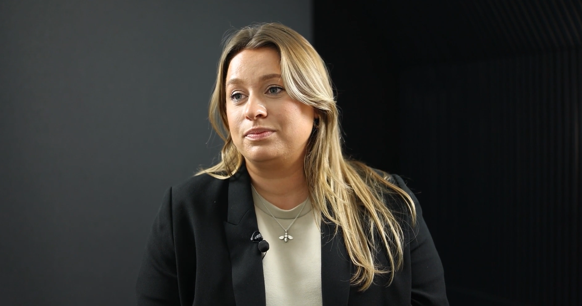 A woman with long blonde hair, wearing a black blazer over a light top, sitting in front of a dark background, looking off to the side.