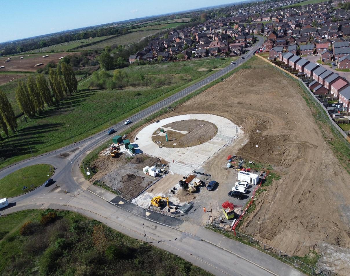 An aerial view of a construction site featuring a circular concrete foundation for a roundabout, surrounded by cleared land and vehicles. In the background, residential houses are visible alongside green fields.