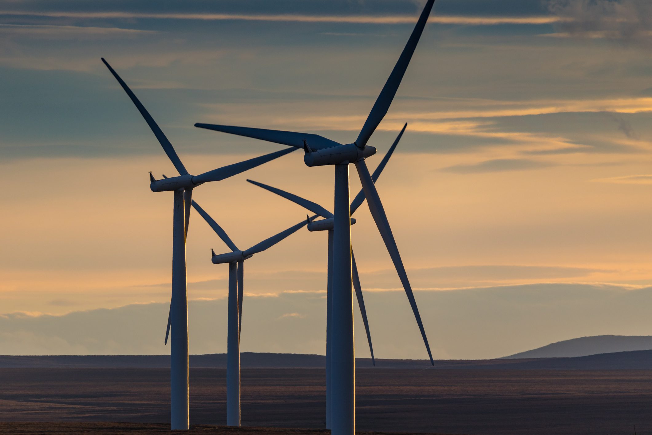 Several wind turbines on a landscape during sunset, with the sky displaying hues of orange and blue. The turbines are oriented slightly in different directions, highlighting their blades against the evening light.