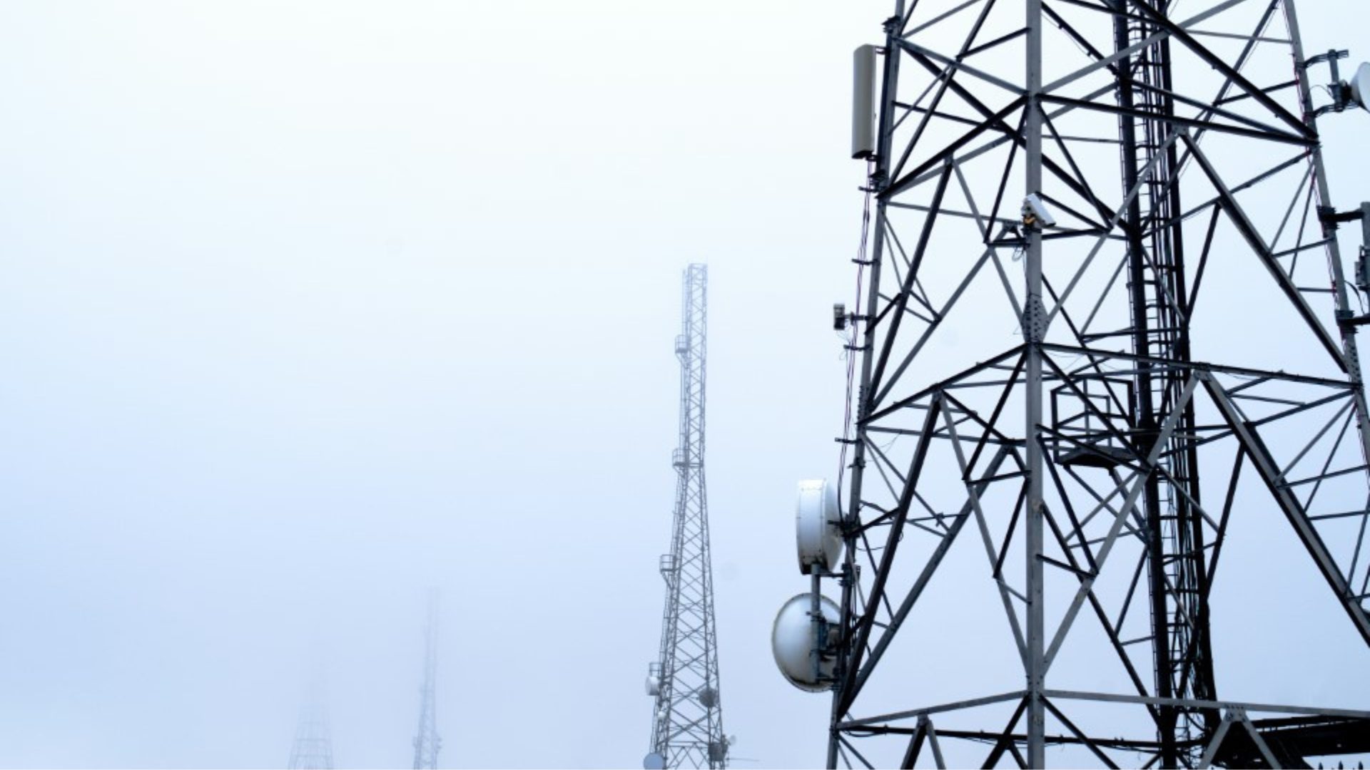Several telecommunications towers with a foggy background, showing metal structures and antennas against a light gray sky.