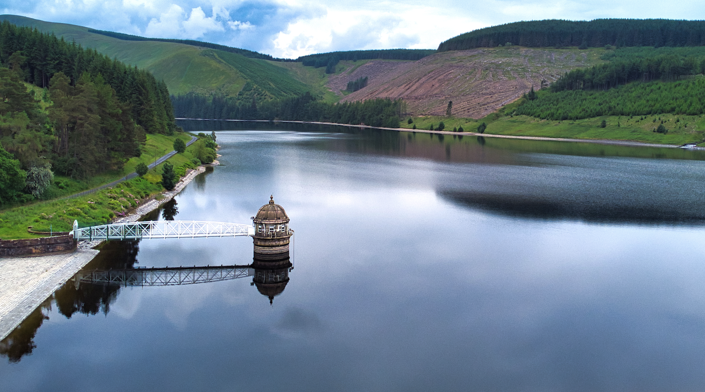 A calm body of water surrounded by lush green hills and forests, featuring a stone jetty with a gazebo. The sky is partly cloudy, reflecting in the water's surface.
