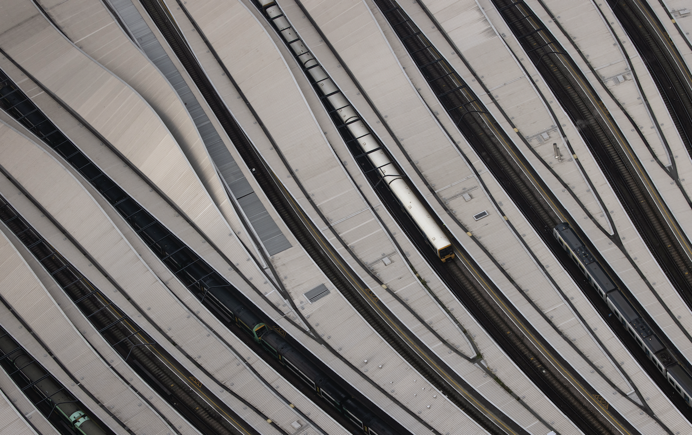 An aerial view of a train station with curved, overlapping roofs and multiple railway tracks. A single train is seen on the tracks amidst the abstract lines of the station's architecture.