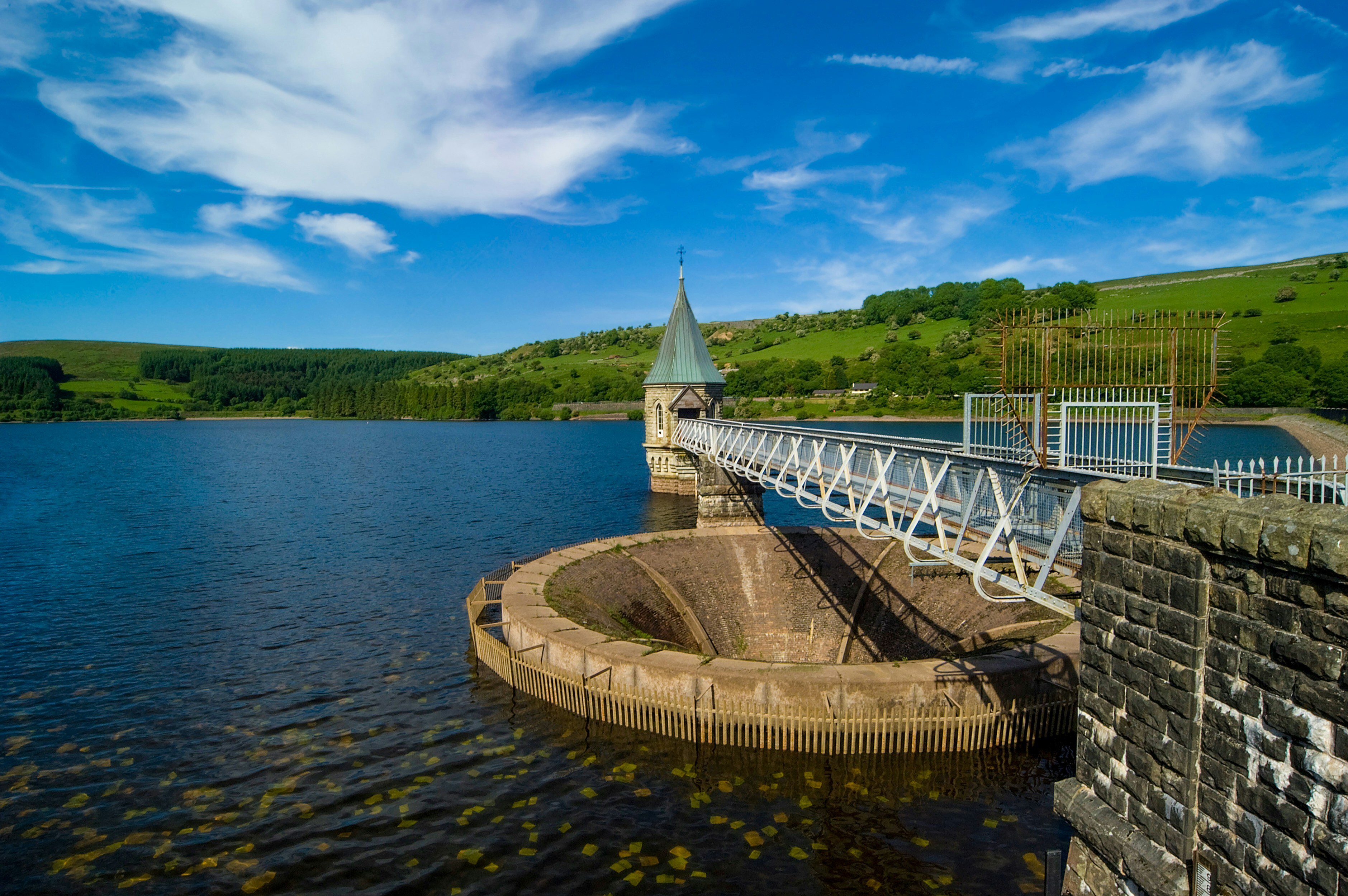 A scenic view of a reservoir with a stone tower and a bridge extending across the water. Lush green hills are visible in the background, while fluffy clouds float in a blue sky.