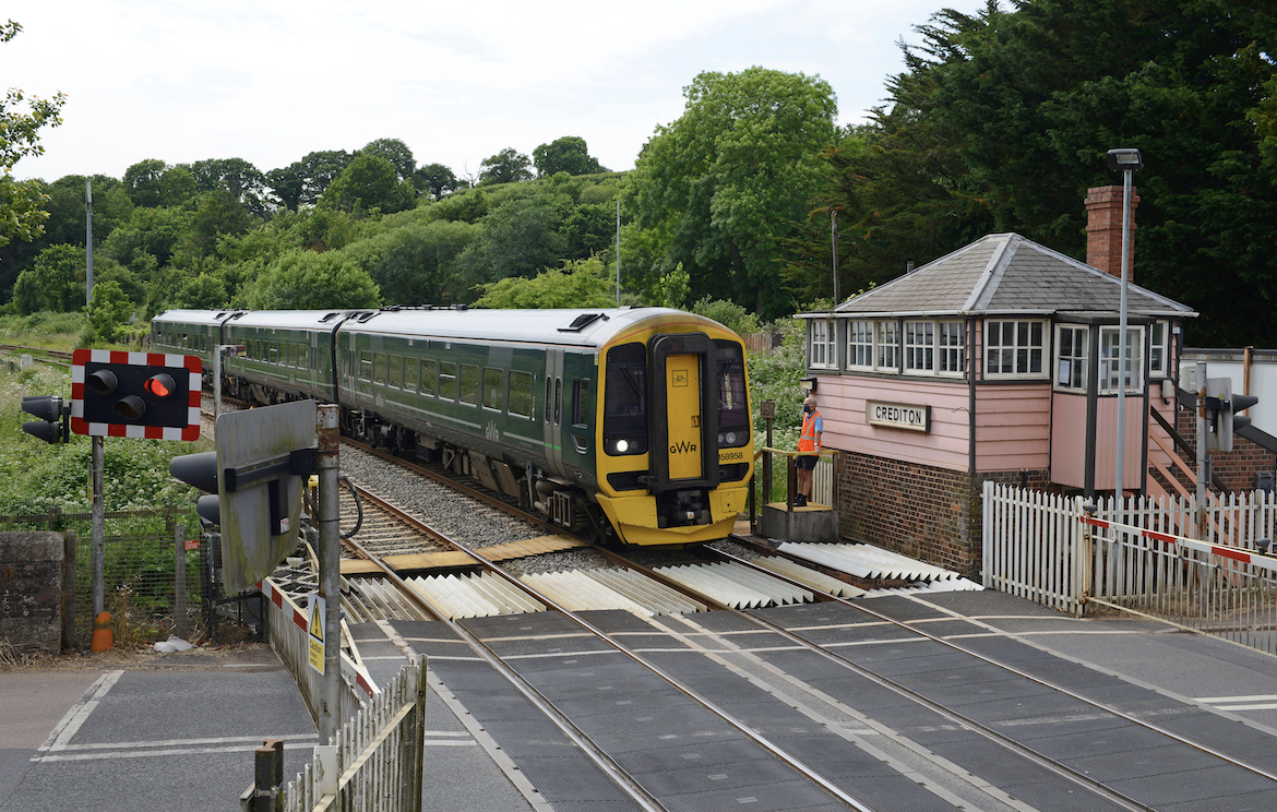 A green commuter train passing through a railway crossing, alongside a signal box labeled "Christon." The area is surrounded by trees, with safety barriers lowered at the crossing.