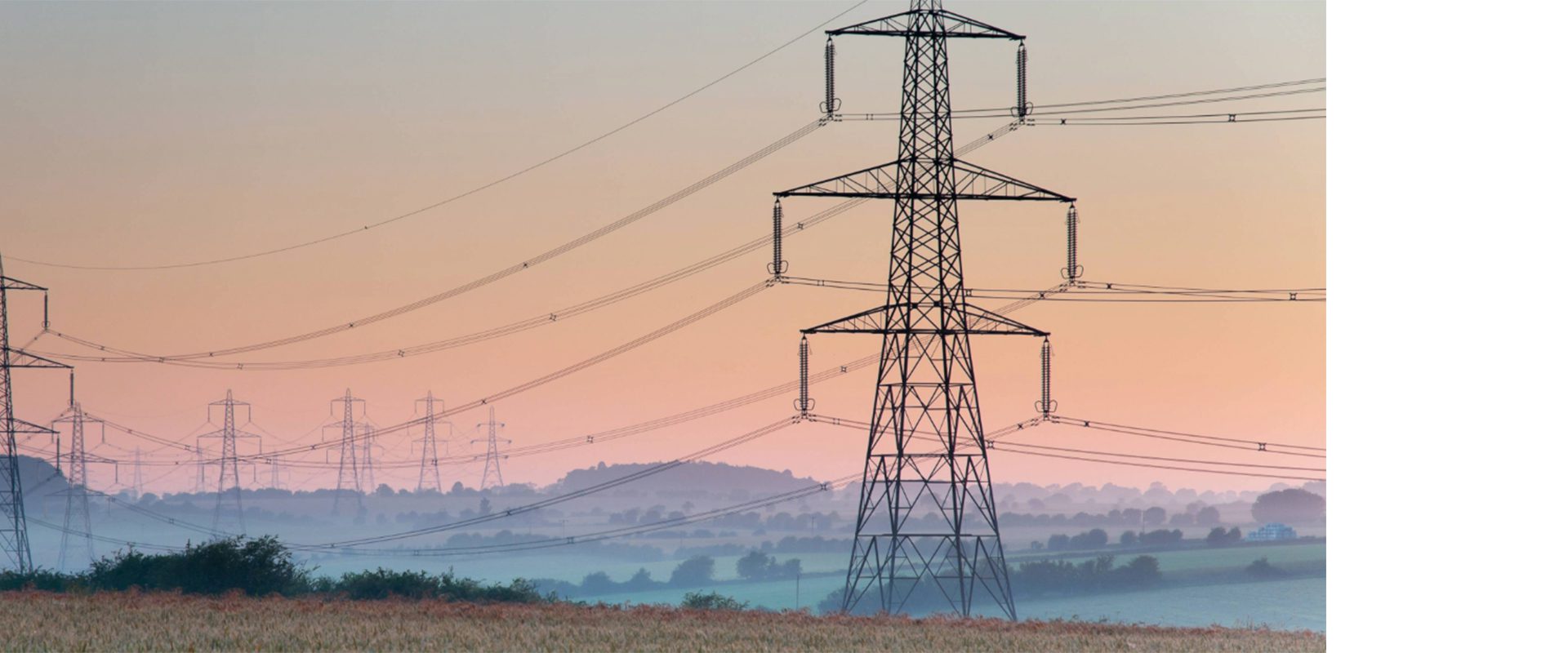 Power transmission towers standing in a rural landscape during early morning, with a soft gradient sky transitioning from purple to orange. Fields stretch in the foreground, and more towers are visible in the distance.