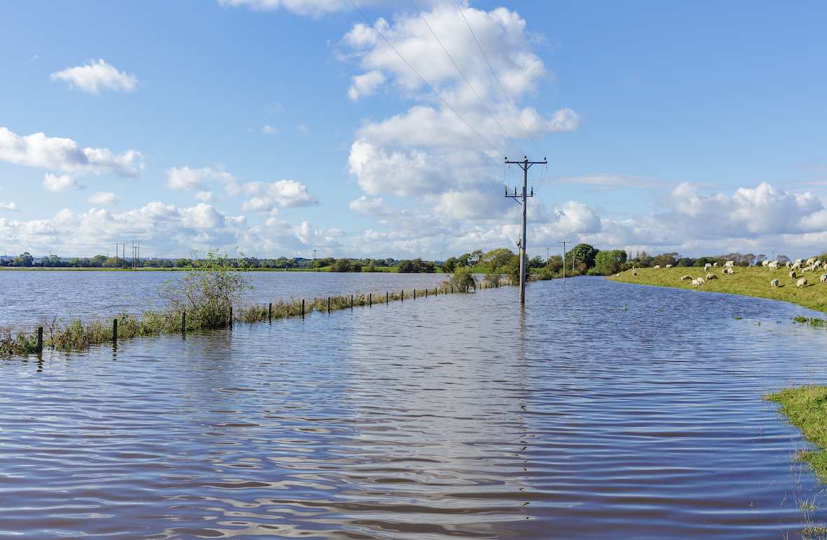 A flooded landscape featuring still water covering fields and a power pole standing amidst the water. Green grass and a fence line are visible along the edge, with clouds scattered in a blue sky.