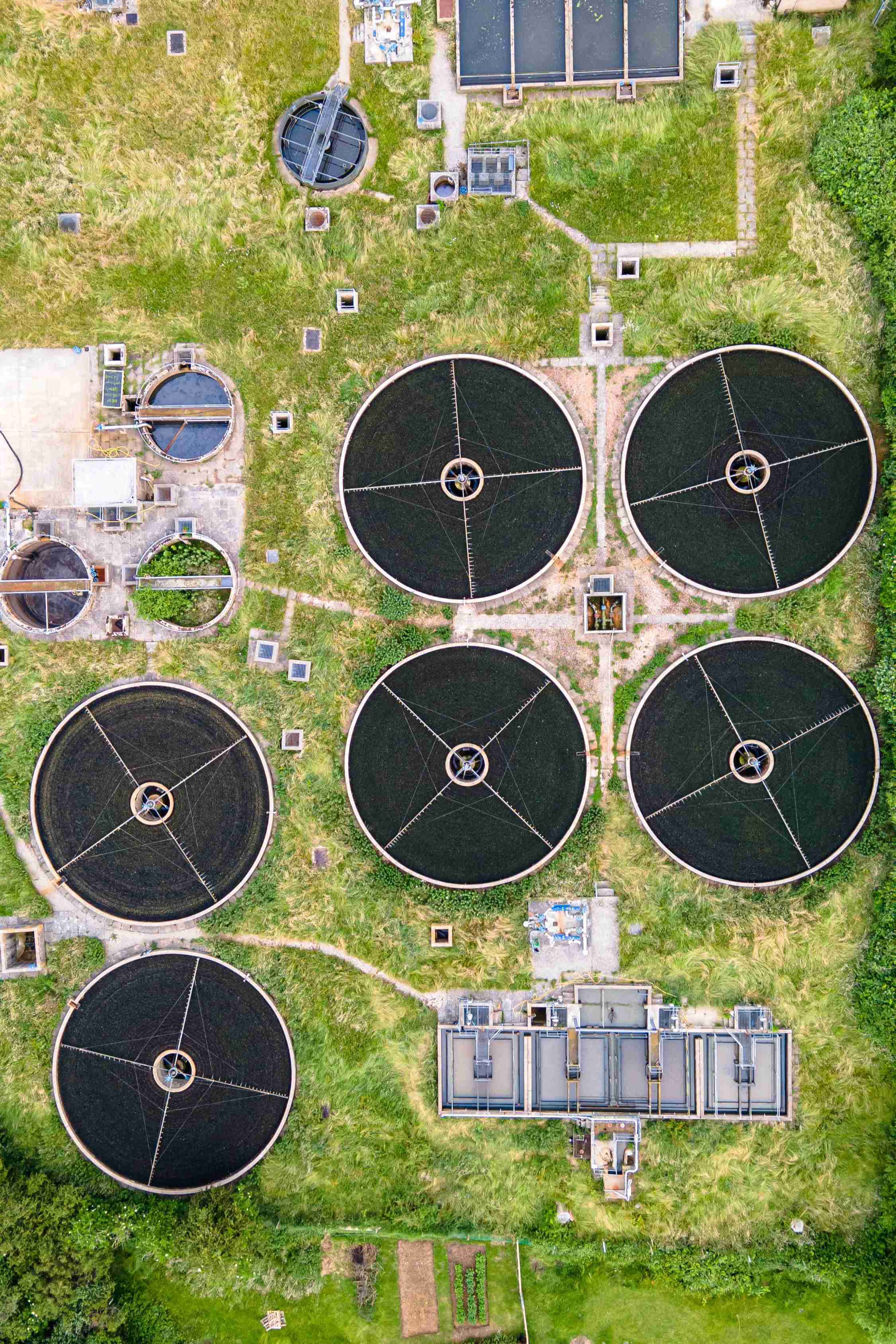 An aerial view of a wastewater treatment facility featuring several large circular tanks surrounded by green grass and buildings. The tanks are dark in color and arranged in a pattern.