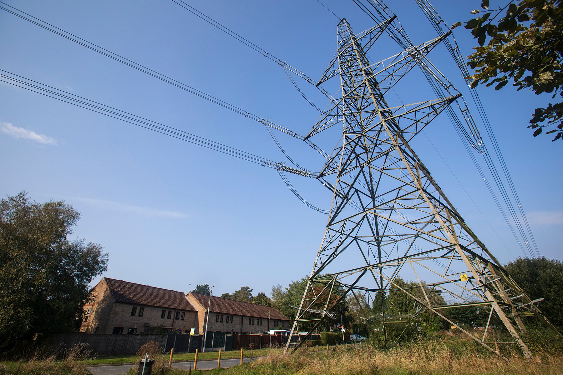 A tall electricity pylon stands beside a residential building with a brown roof, surrounded by greenery. Blue sky and a few clouds are visible in the background.