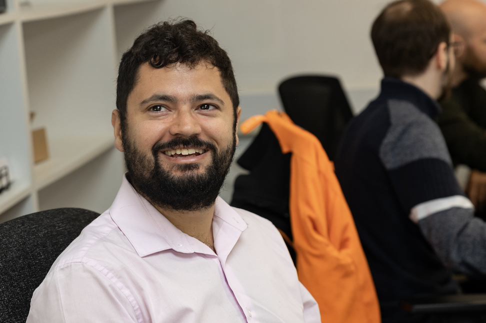 A man with a beard wearing a pink shirt, smiling while sitting at a desk with a black chair. In the background, a person in a green shirt and another in a gray sweater are working at separate desks.
