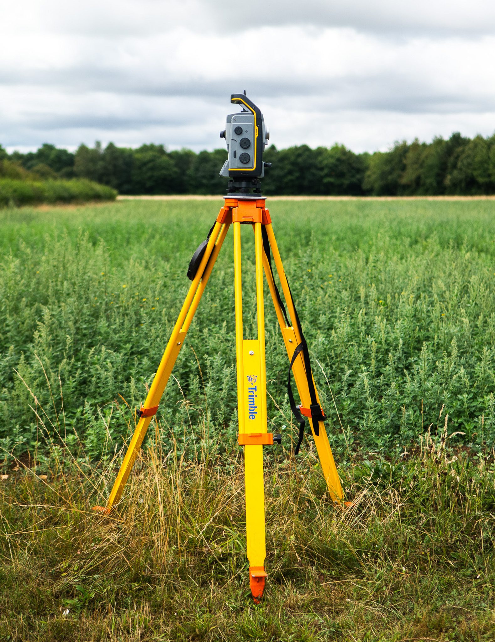 A surveying instrument mounted on a yellow tripod, positioned in a green field with trees in the background under a cloudy sky.