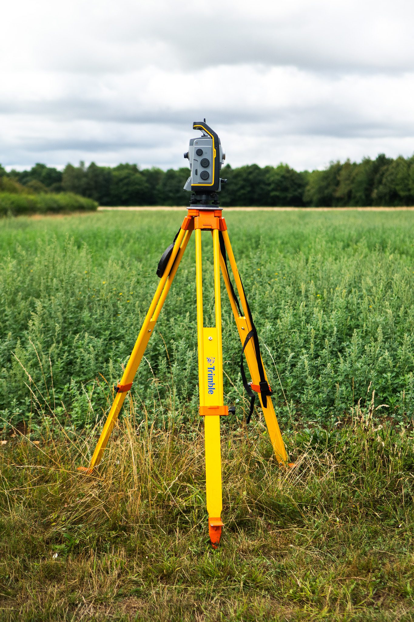 A surveying instrument mounted on a yellow tripod in a green field, with trees in the background under a cloudy sky.