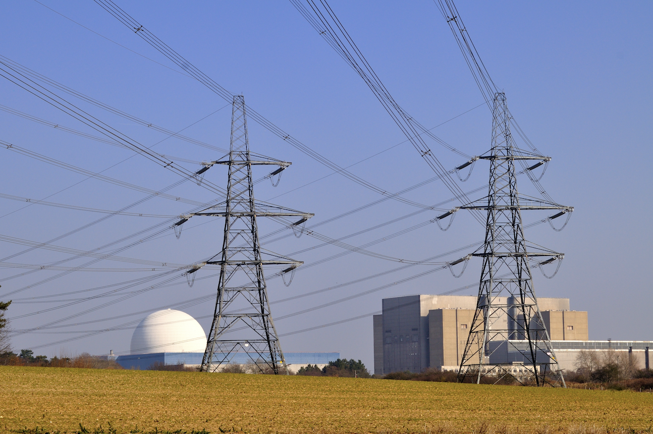 Electricity pylons in a field with a nuclear power plant in the background. The plant features a large dome and a modern industrial building against a clear blue sky.
