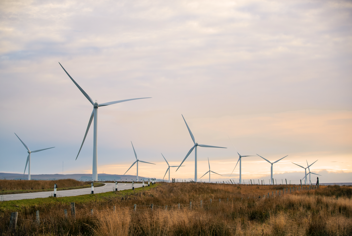 A landscape featuring multiple wind turbines beside a pathway, with grassy fields and a cloudy sky in the background. The scene emphasizes renewable energy and open spaces.