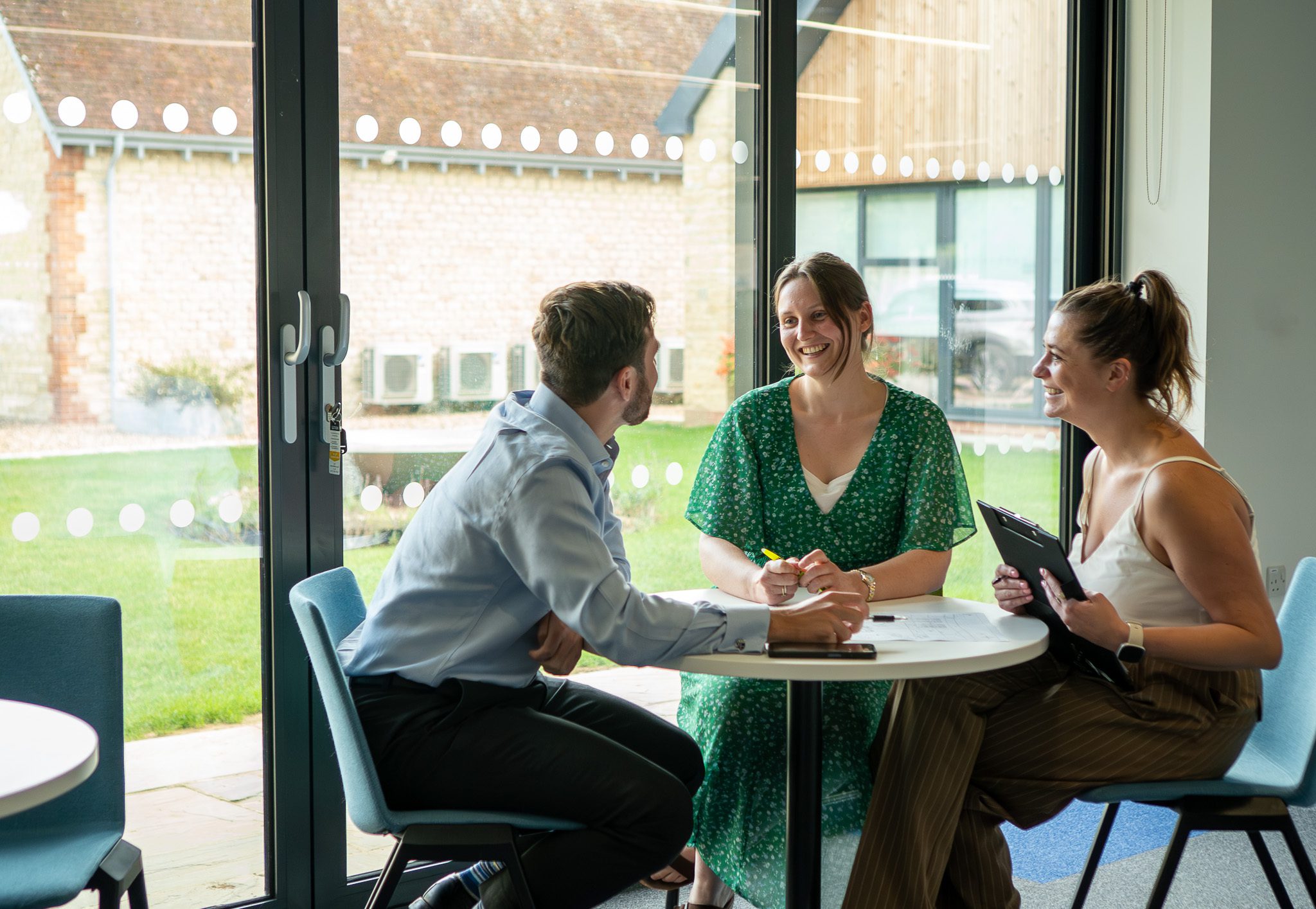 Three professionals engaged in discussion around a table in a bright office space, with large windows showing a grassy outdoor area. One person is writing notes, while another holds a tablet.