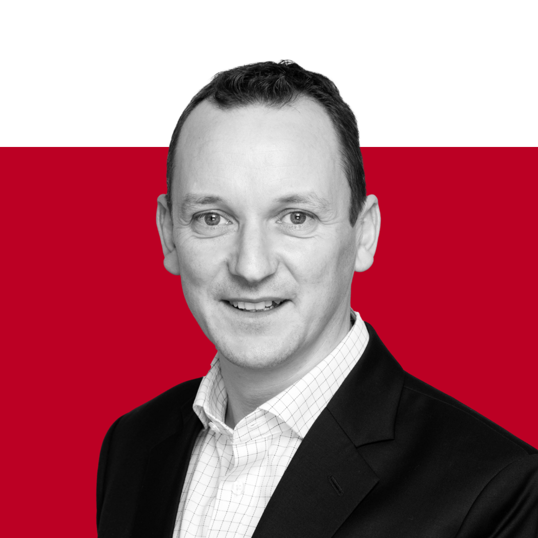 A professional headshot of a man in a black blazer and white shirt against a red background. He is smiling directly at the camera.