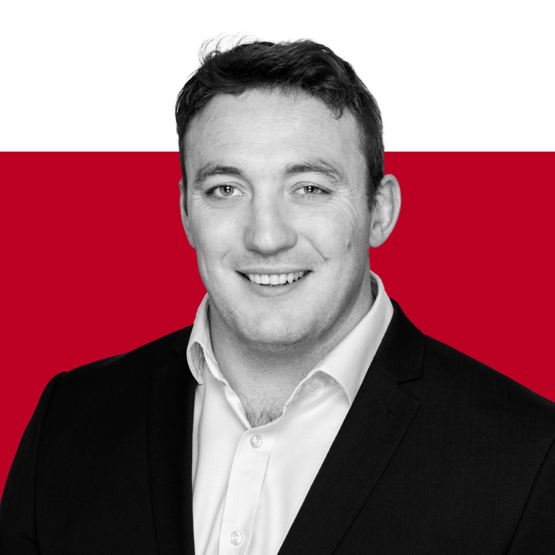 A smiling man in a black suit and white shirt, with short dark hair, against a red background.