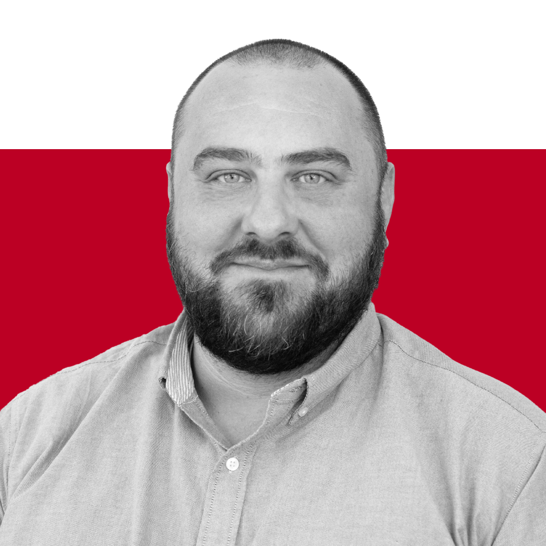 A man with a beard wearing a light-colored button-up shirt, standing in front of a solid red background.