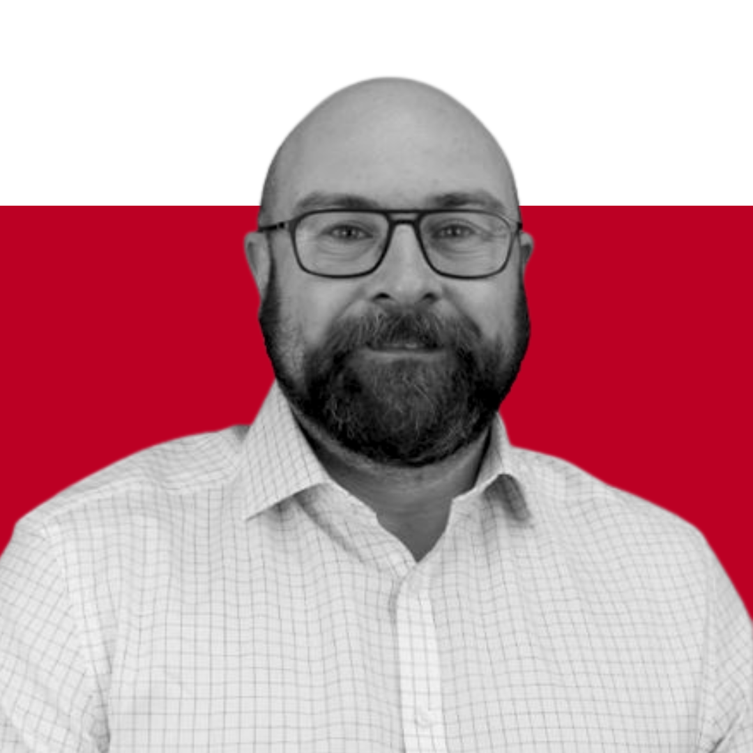 A man with a beard wearing glasses and a light checkered shirt, set against a red background. He is looking directly at the camera.