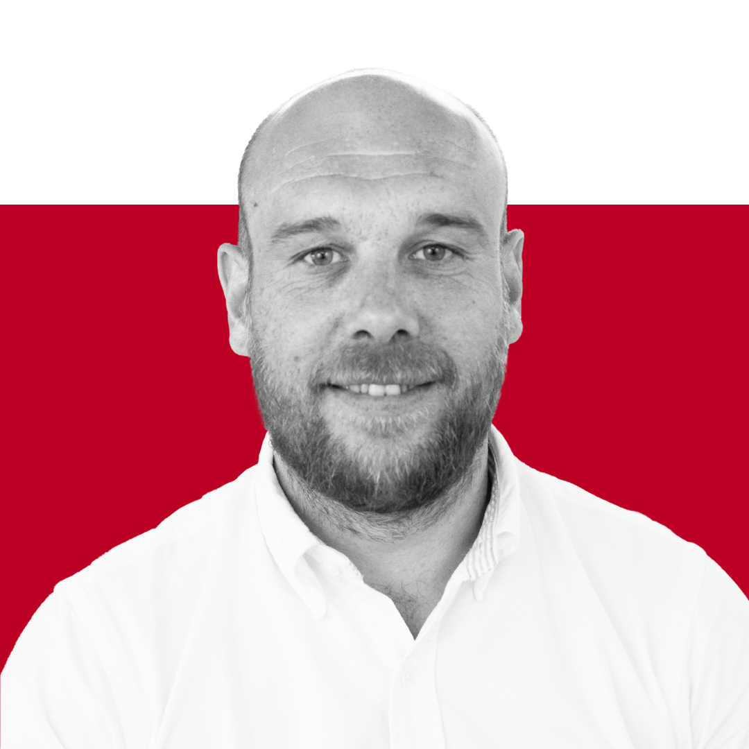 A smiling man with a shaved head and a beard, wearing a white collared shirt, is photographed against a red and black background.