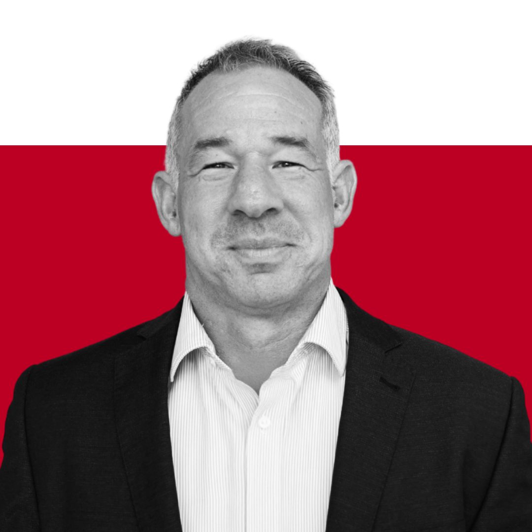 A man in a suit smiling in front of a red and white background, with his face in black and white. He appears confident and approachable.