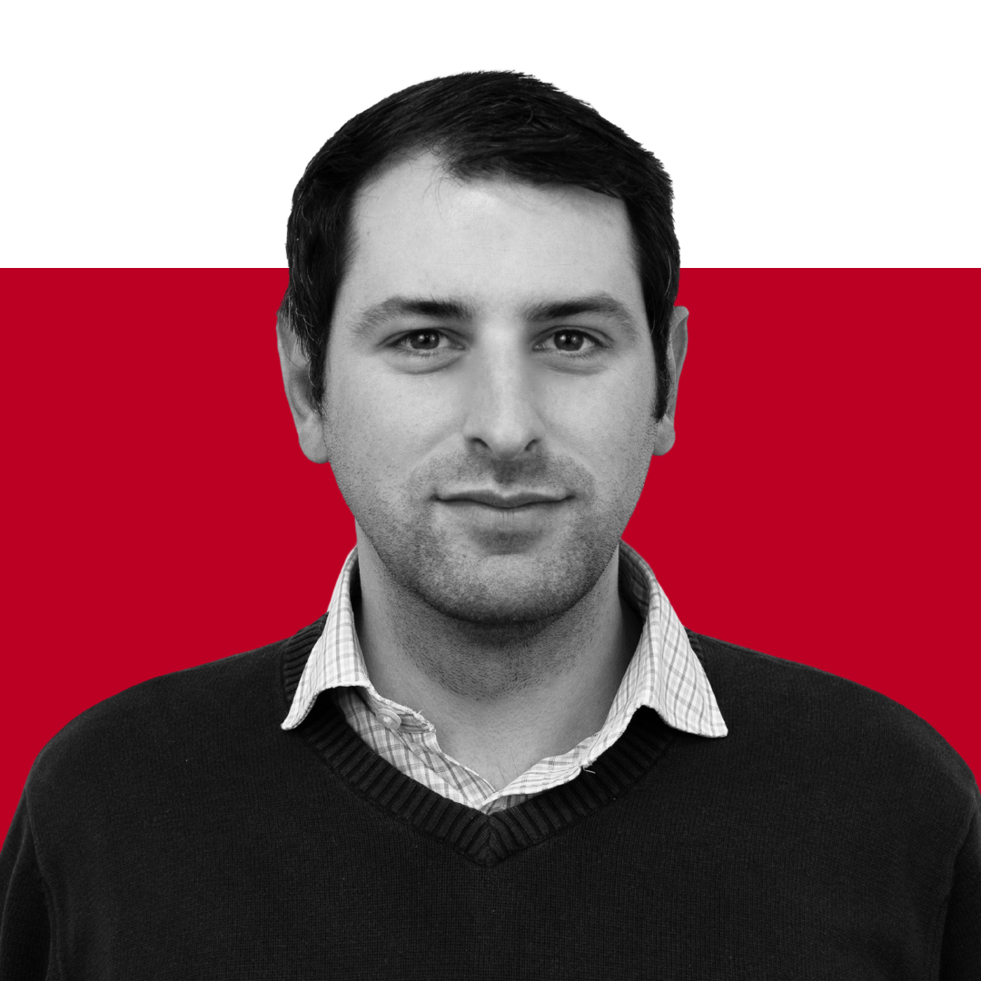 A man with dark hair wearing a dark sweater over a collared shirt, posed against a background split between black and red. He has a neutral expression and is facing the camera.