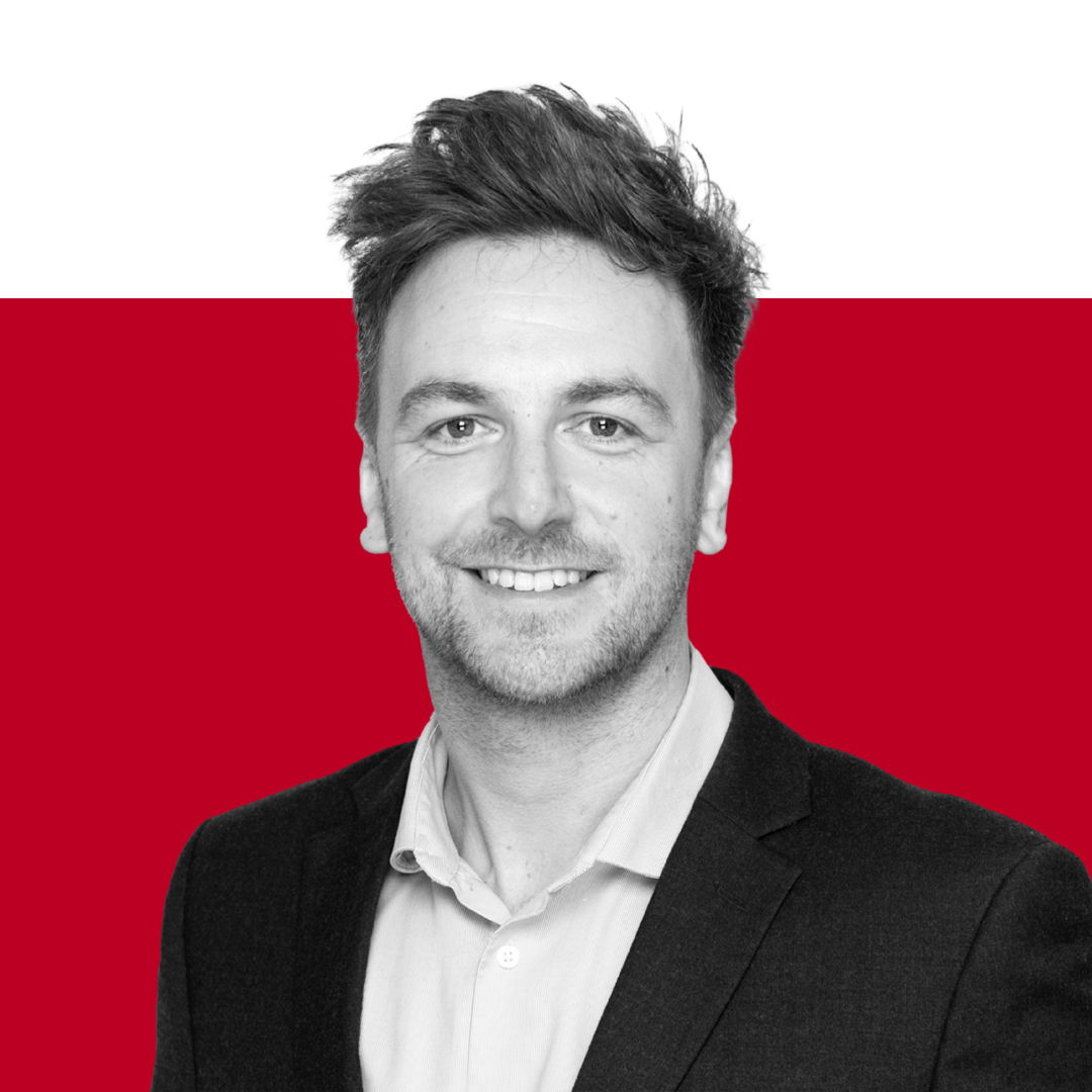 A professional headshot of a man smiling, wearing a black suit and a light-colored shirt, with a red and black background.