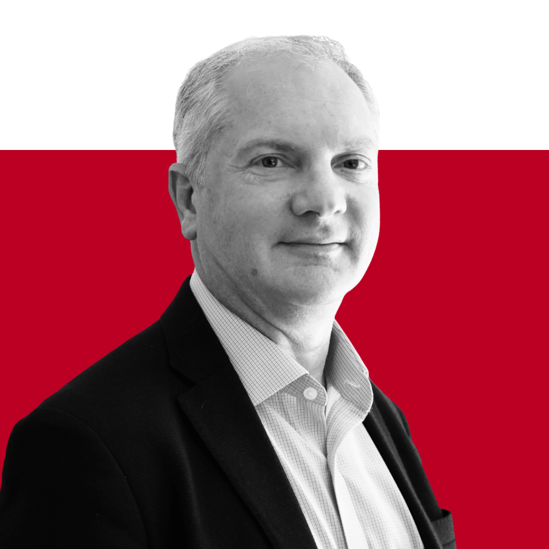 A man with short, light-colored hair and wearing a black blazer over a collared shirt, posing against a red background. He is looking slightly towards the camera.