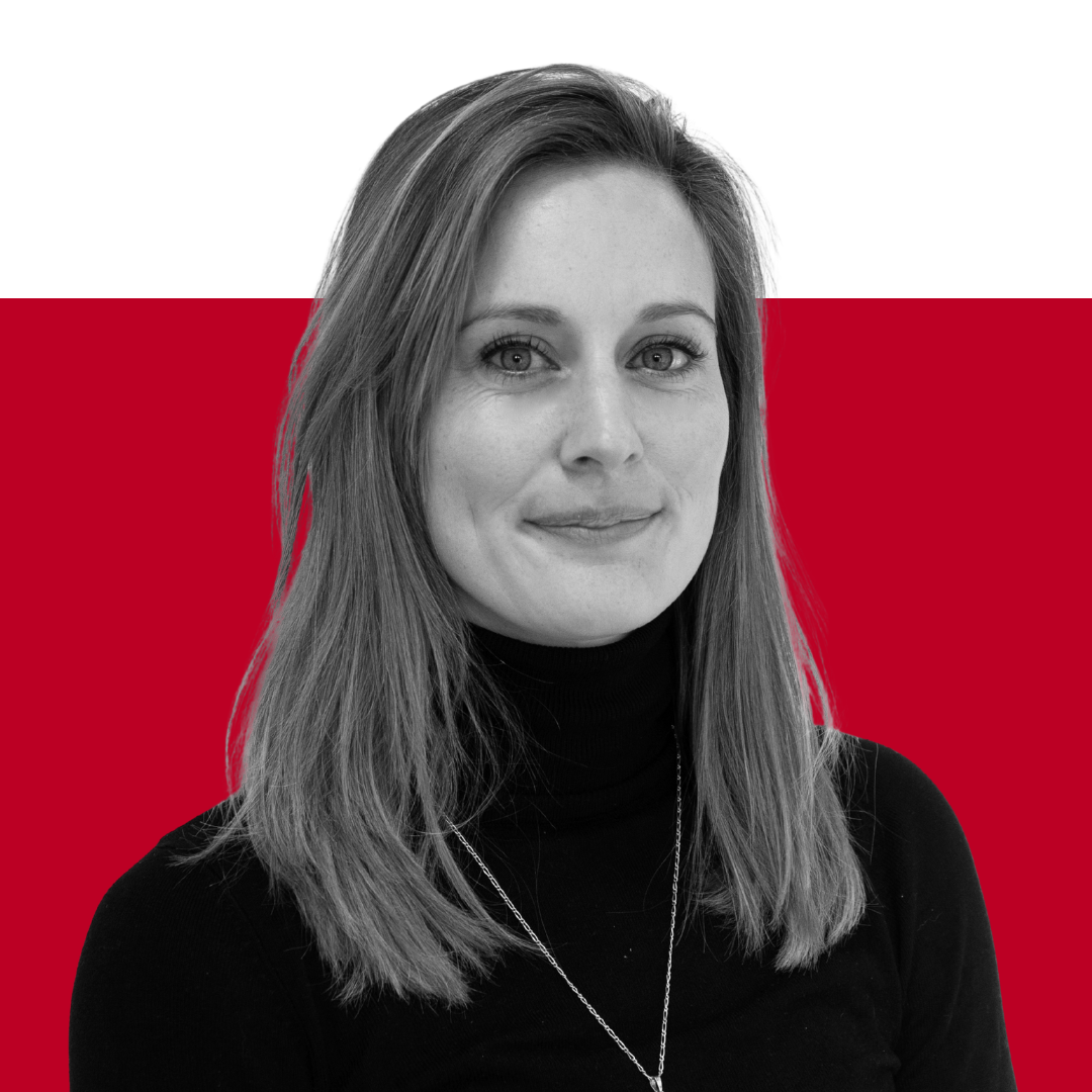 A woman with long brown hair wearing a black turtleneck sweater, smiling while facing the camera. The background features a solid black upper section and a solid red lower section.