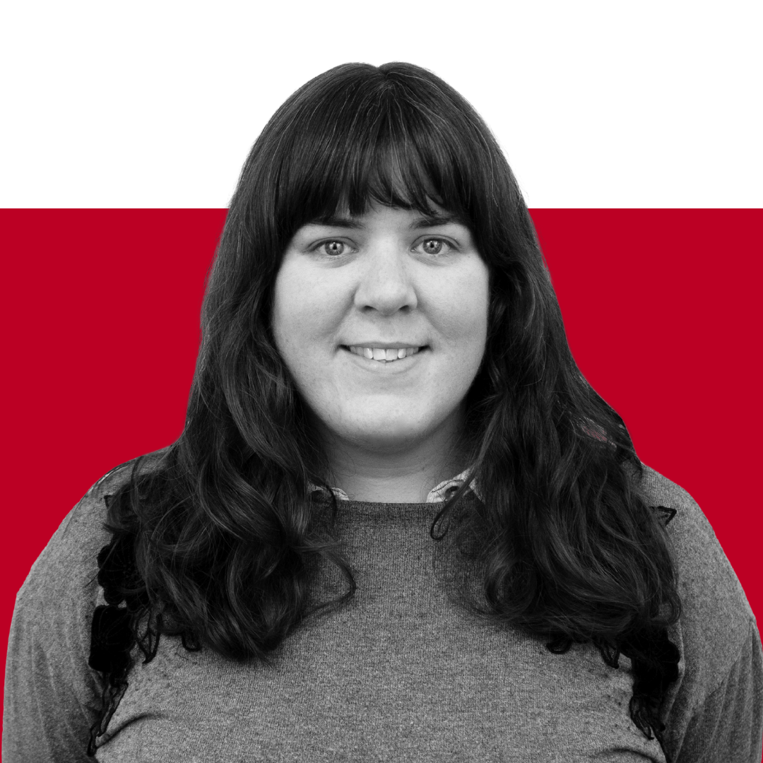 A woman with long dark hair and a gray top smiling in front of a red and black background.