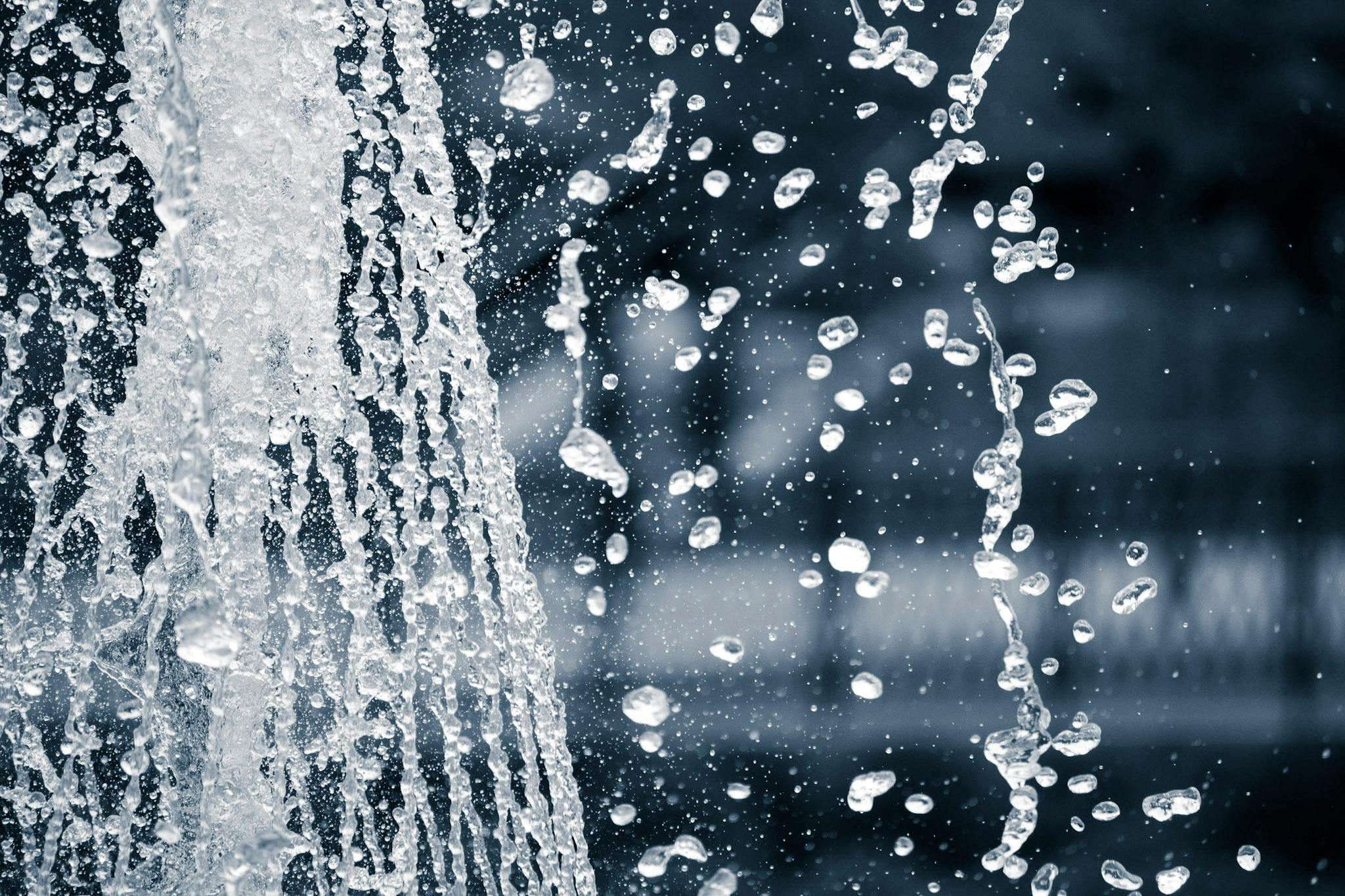 Close-up of water droplets splashing from a fountain against a blurred background. The image captures the motion and texture of the water as it cascades.