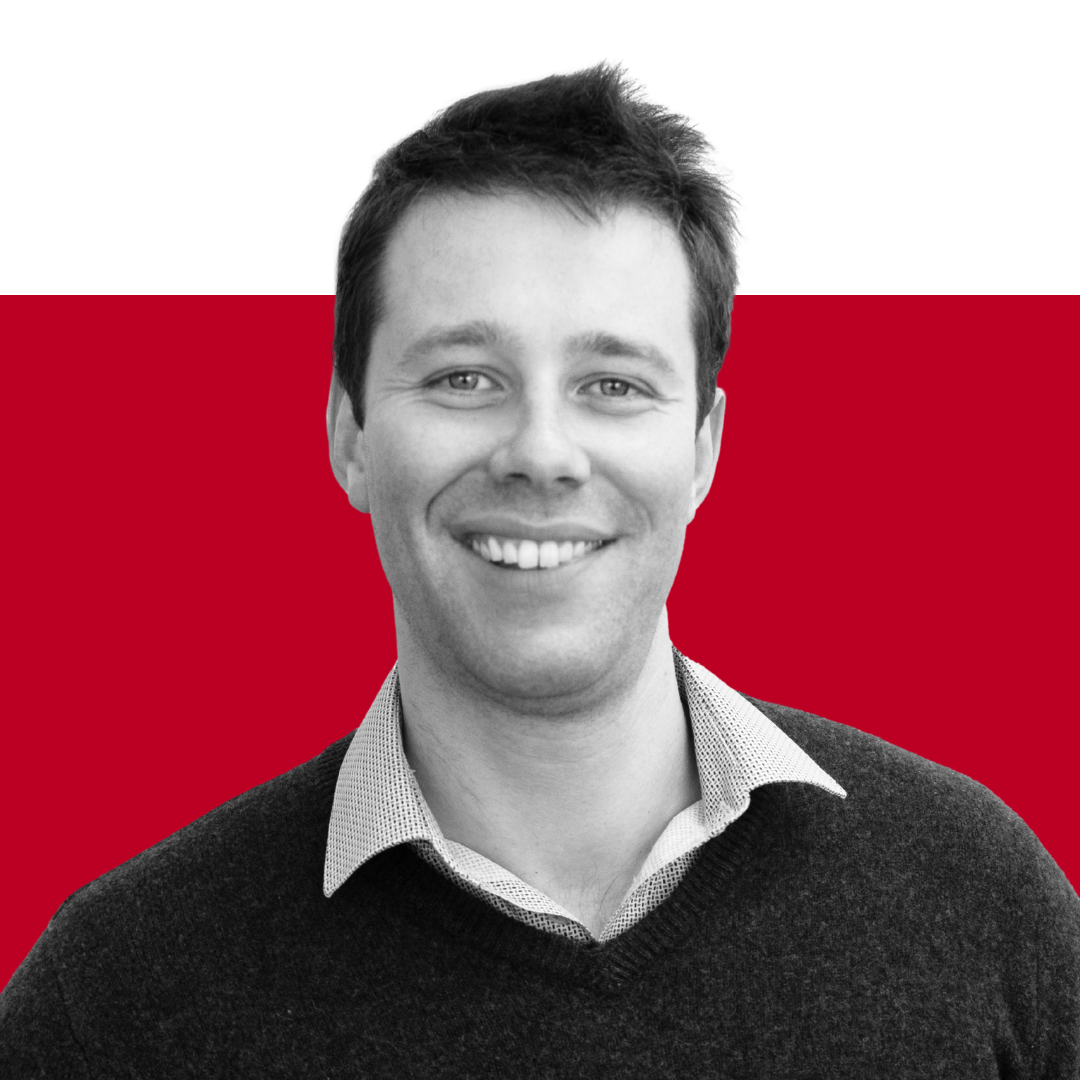 A smiling man with short dark hair, wearing a light gray collared shirt and black sweater, set against a black and red background.